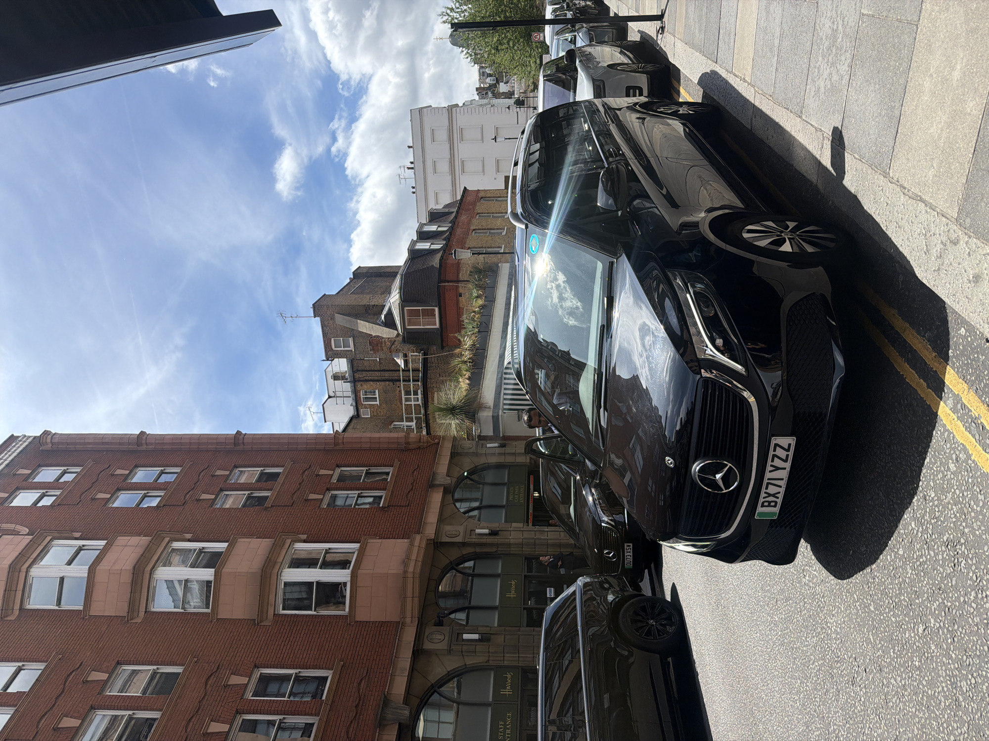 The image shows a street scene in London, United Kingdom, with several vehicles parked along the curb. In the foreground, a black Mercedes-Benz V-Class van is prominently featured. Its license plate reads "BX71 YZZ". To its left, a black SUV is partially visible. Further down the street, a white car and other vehicles are parked in a line. The background consists of buildings, including a red brick building with arched entrances marked "Harrods" and a white building. The sky is partly cloudy. A man in a suit is standing near the Harrods entrance, looking towards the vehicles. The street is paved with asphalt and lined with a sidewalk made of stone slabs. The overall impression is a bustling urban environment on a sunny day.