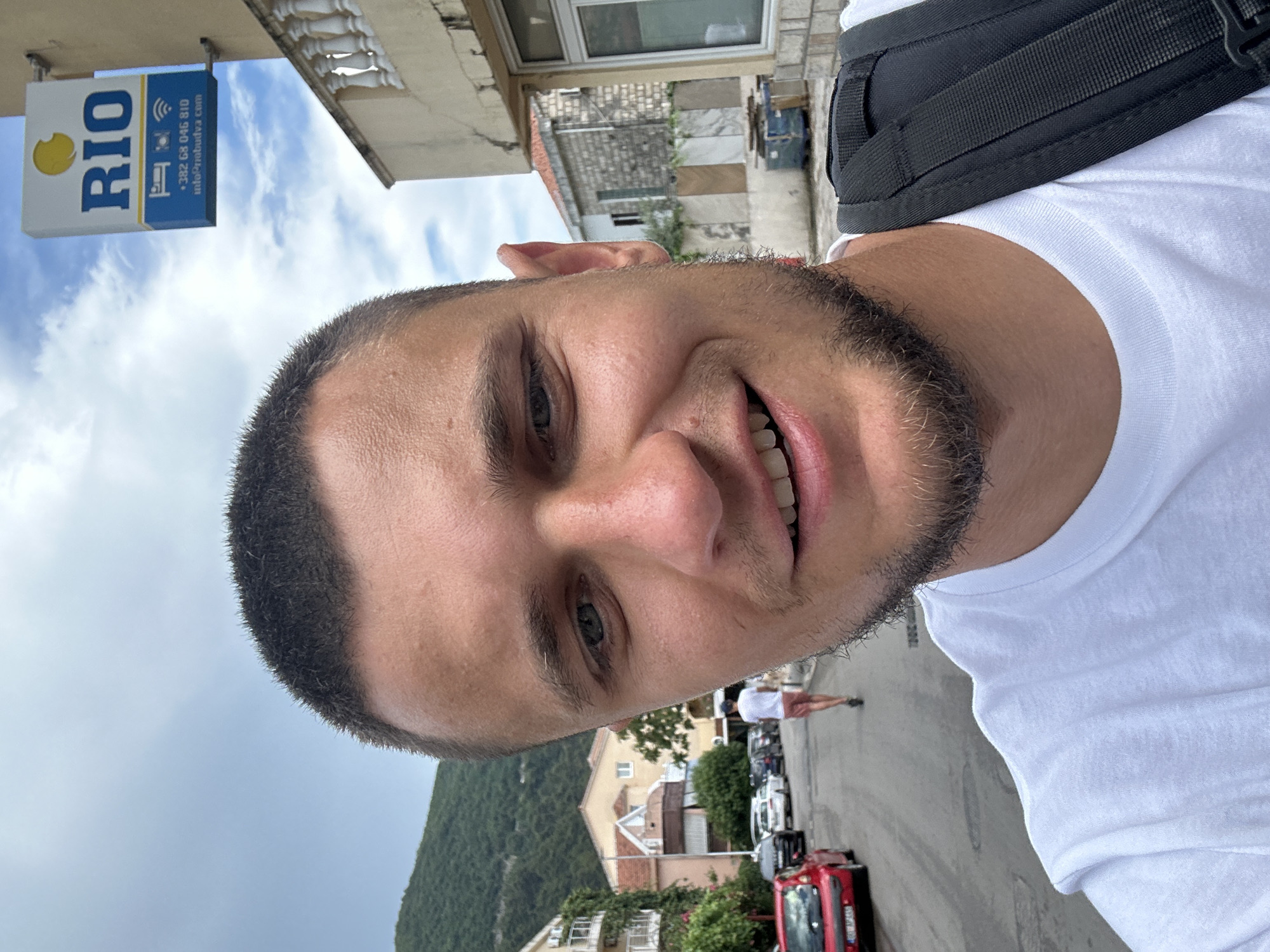 In Budva, Montenegro, a man with short dark hair and a light beard is captured in a close-up selfie. He is wearing a white t-shirt and has a black backpack strap visible over his right shoulder. Behind him, a street is lined with parked cars, including a red hatchback on the left. Further down the street, a person in a white t-shirt and red shorts is walking away from the camera. Buildings with balconies and tiled roofs flank the street, and a lush green mountain rises in the background under a cloudy sky. A sign for "RIO" with contact information and icons indicating accommodation, a phone, and Wi-Fi is mounted on a building to the right of the frame.
