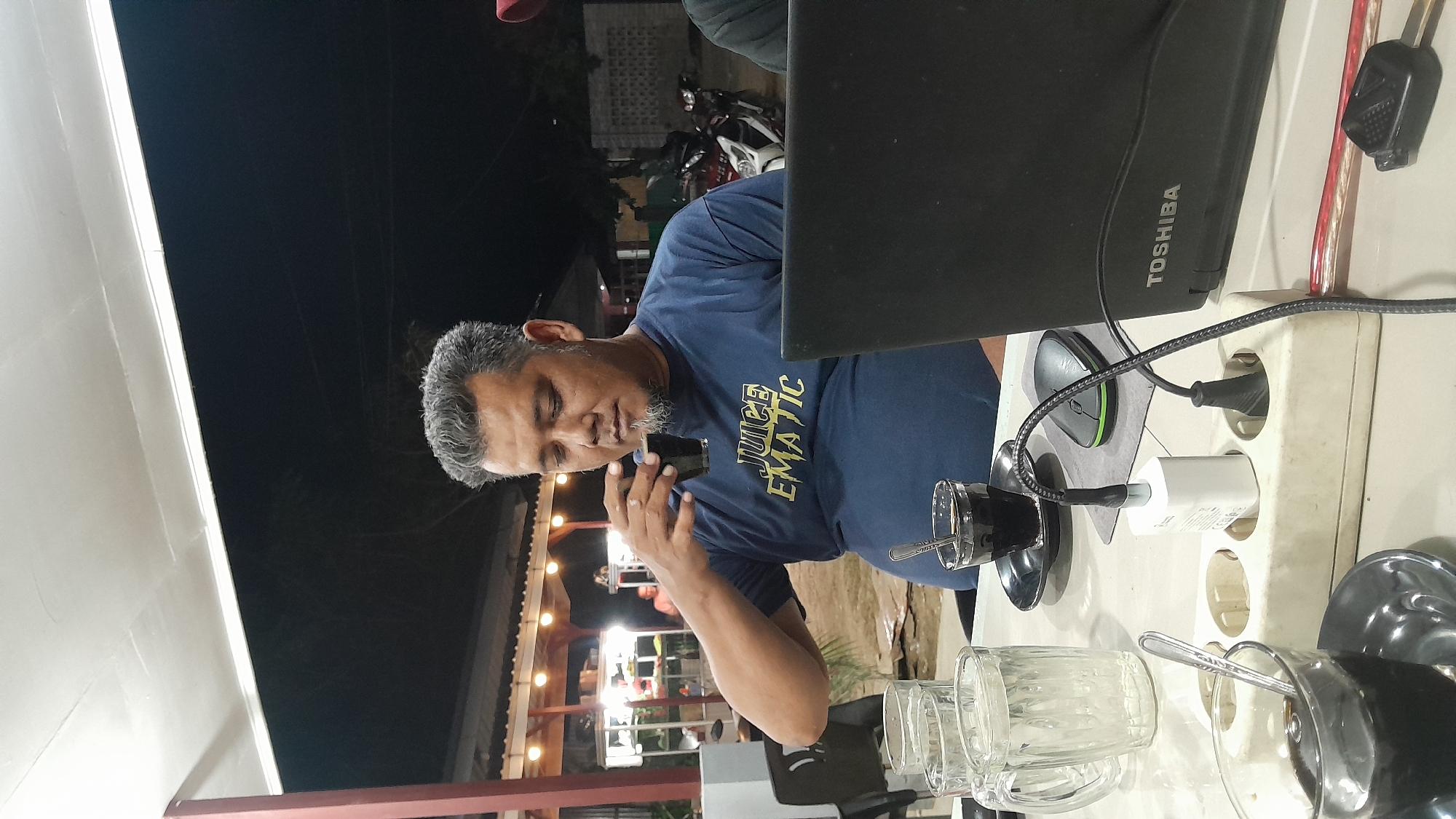 A man with graying hair and a beard is seated at an outdoor table. He wears a blue t-shirt with the words "Juice EMATIC" printed on it. He is holding a small glass to his lips. On the table, there is a black Toshiba laptop, a mouse, and a white power strip with several plugs inserted. There are also multiple glasses and cups, some with spoons in them. In the background, there is a building with lights and a motorcycle. The scene is in Lhokseumawe, Indonesia. The time is nighttime.