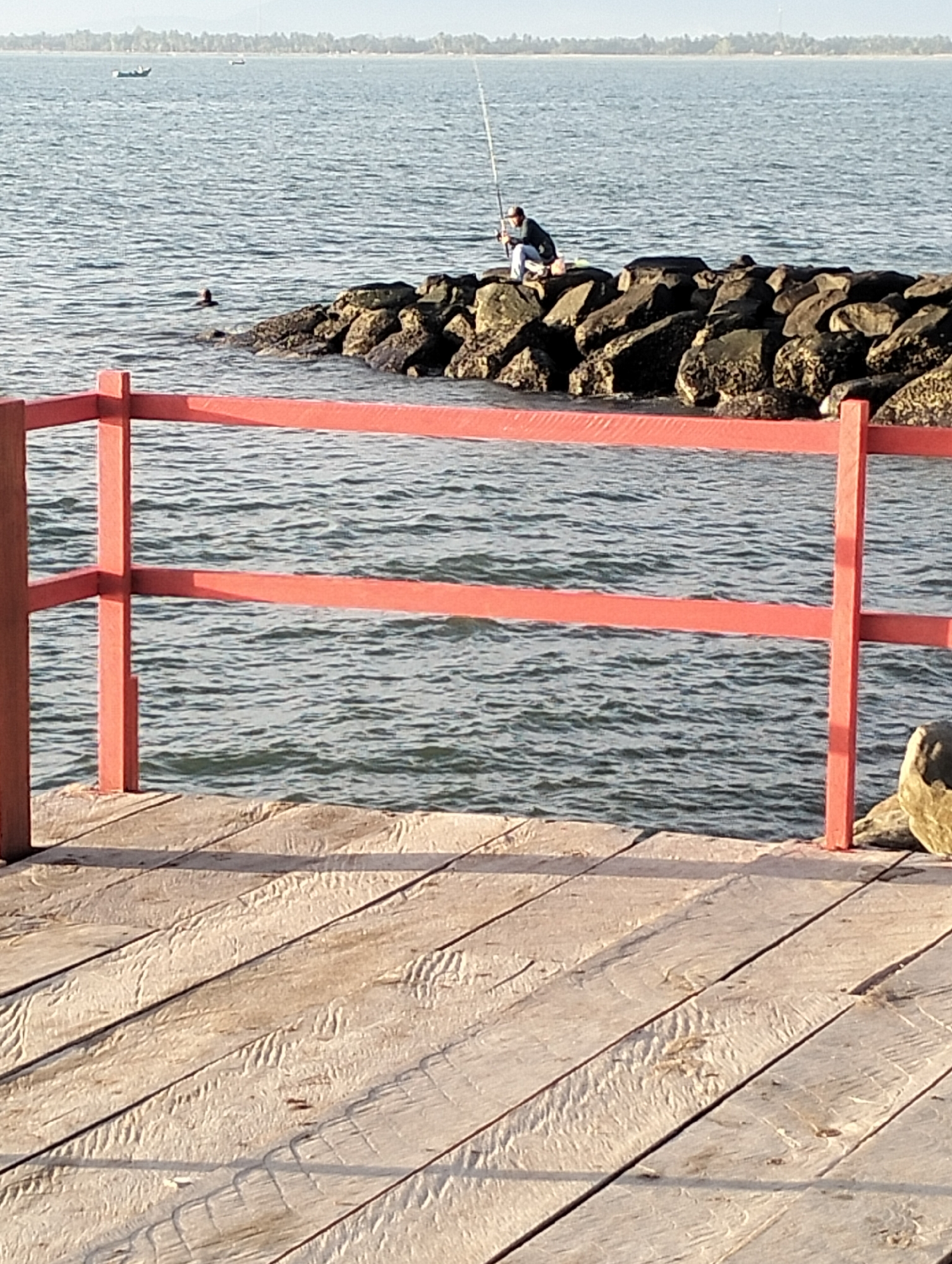 The image depicts a coastal scene at a location in Lhokseumawe, Indonesia. In the foreground, a wooden pier with a red railing runs horizontally across the lower half of the frame. The pier's surface is made of weathered wooden planks. Beyond the pier, a body of water, likely the sea, stretches to the horizon. A breakwater constructed of large rocks extends into the water. On the breakwater, a person is visible, seated and holding a fishing rod, engaged in fishing. Further in the water, another person is seen swimming. In the distance, a thin treeline borders the coastline, and a small boat is present on the water. The sky is clear and bright.