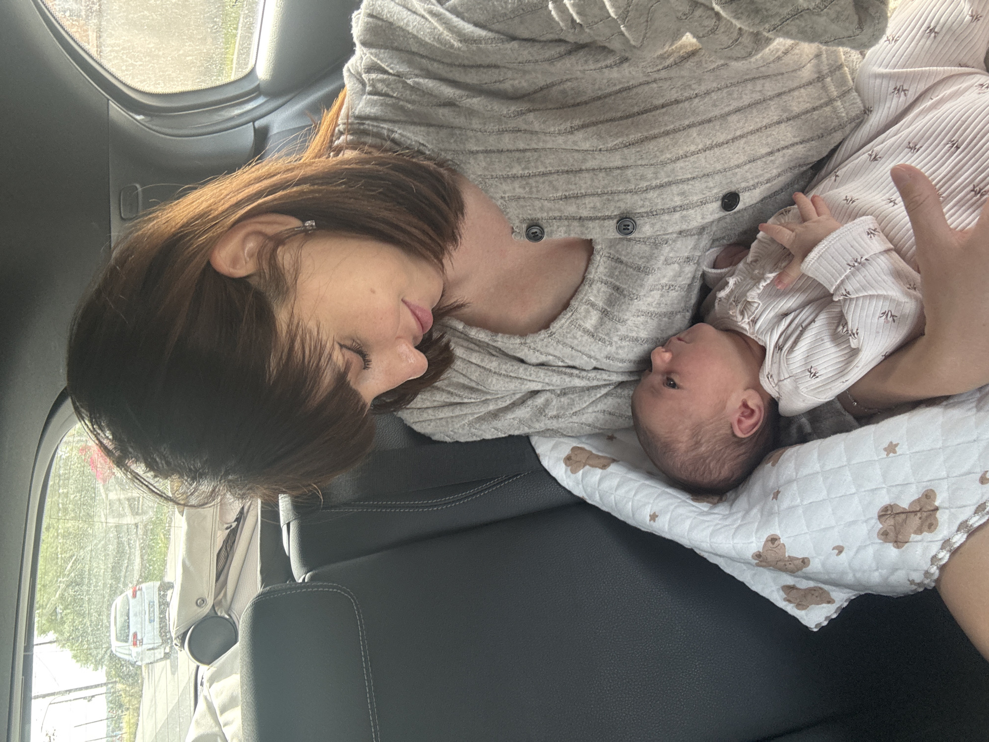 A woman with brown hair and fair skin lies on a black car seat, cradling a baby wrapped in a blanket with teddy bear patterns. The woman wears a gray, horizontally-striped, buttoned shirt. The baby wears a patterned, white onesie. The view through the car window shows a street in Nettuno, Italy, with buildings and a white car. The scene is bathed in soft, diffused light.