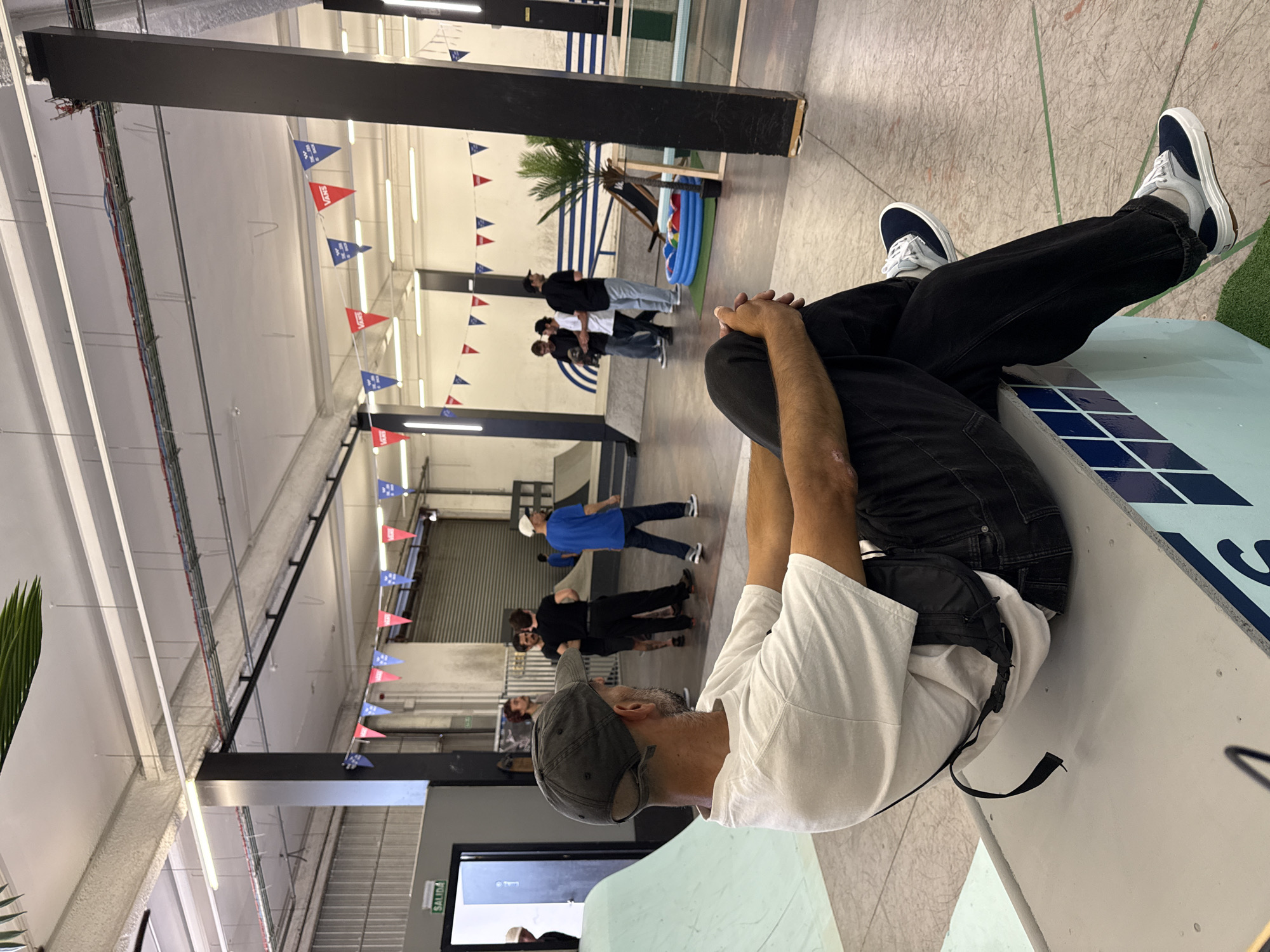 A man in a white t-shirt, black jeans, and dark sneakers sits on the edge of a skate ramp indoors. He is wearing a gray baseball cap and has a black messenger bag slung over his shoulder. In the background, several people are standing or walking around the skate park. There are Vans brand pennants hanging from the ceiling, as well as a small swimming pool with a palm tree. The location is L'Hospitalet De Llobregat, Spain.