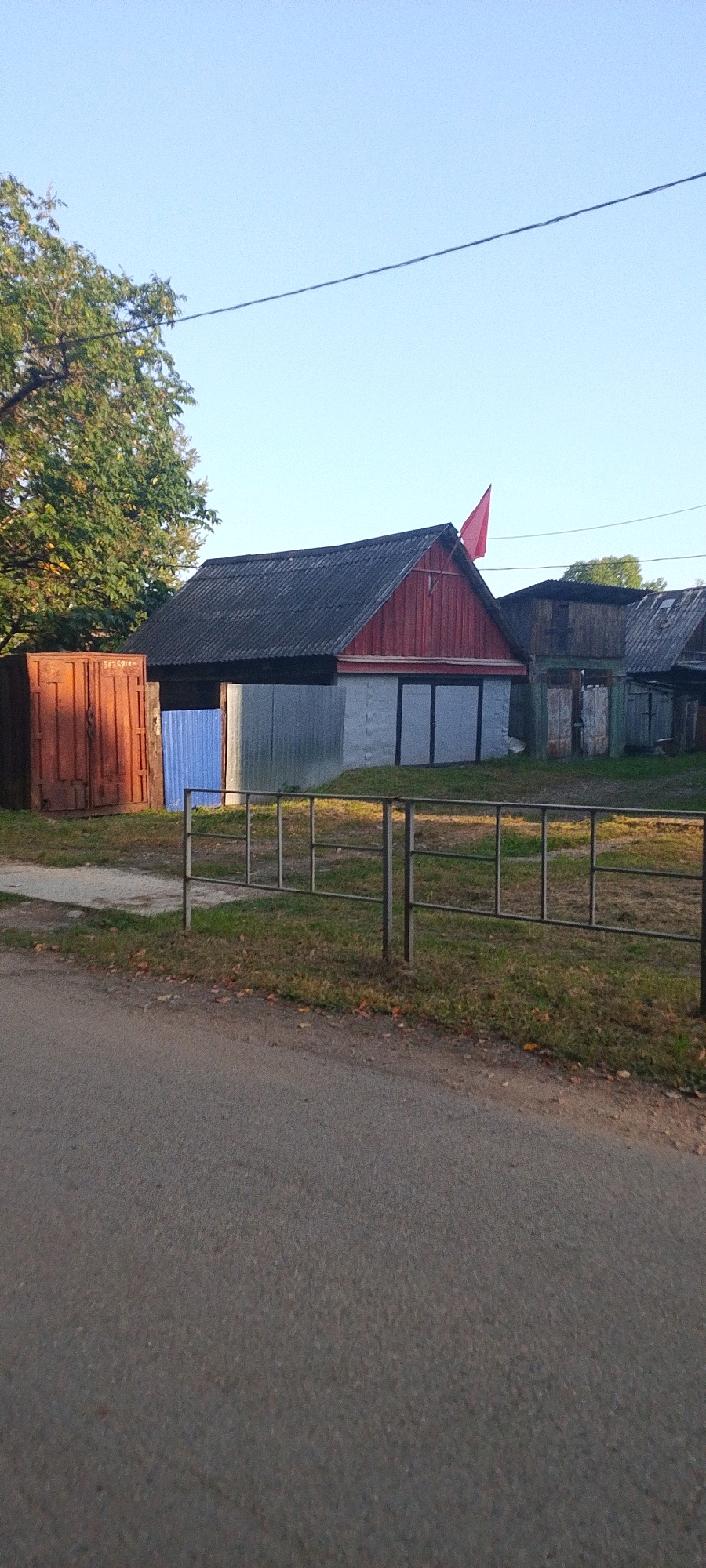The image depicts a rural scene in Bira, Russia, featuring a collection of structures and natural elements. A long, rusty metal container or shed, possibly made of corrugated iron, stands on the left side of the frame. Adjacent to it is a bright blue metal fence or gate.
Dominating the center of the image is a rustic building with a dark, weathered roof, possibly made of corrugated asbestos-cement sheets. The front gable of this building is painted red and features a light-colored facade below, with what appear to be large double doors with dark frames. A bright red flag is prominently displayed on the peak of the red gable, angled towards the left.
To the right of this central structure, another building with a similar dark roof and wooden facade is visible, suggesting a row of utility or storage buildings.
A simple metal railing, composed of vertical posts and horizontal bars, separates the immediate foreground from the area where the buildings are located. The ground in front of the railing is covered with grass and scattered fallen leaves, indicating an autumn or late summer setting. A paved road runs along the bottom of the image, suggesting it is a street or path leading through the area.
A mature tree with green foliage is visible behind the rusty container on the left, and the sky above is a clear, pale blue, with a thin power line stretching horizontally across the upper portion of the frame. The overall impression is one of a functional, perhaps somewhat neglected, rural or industrial outskirts area.
