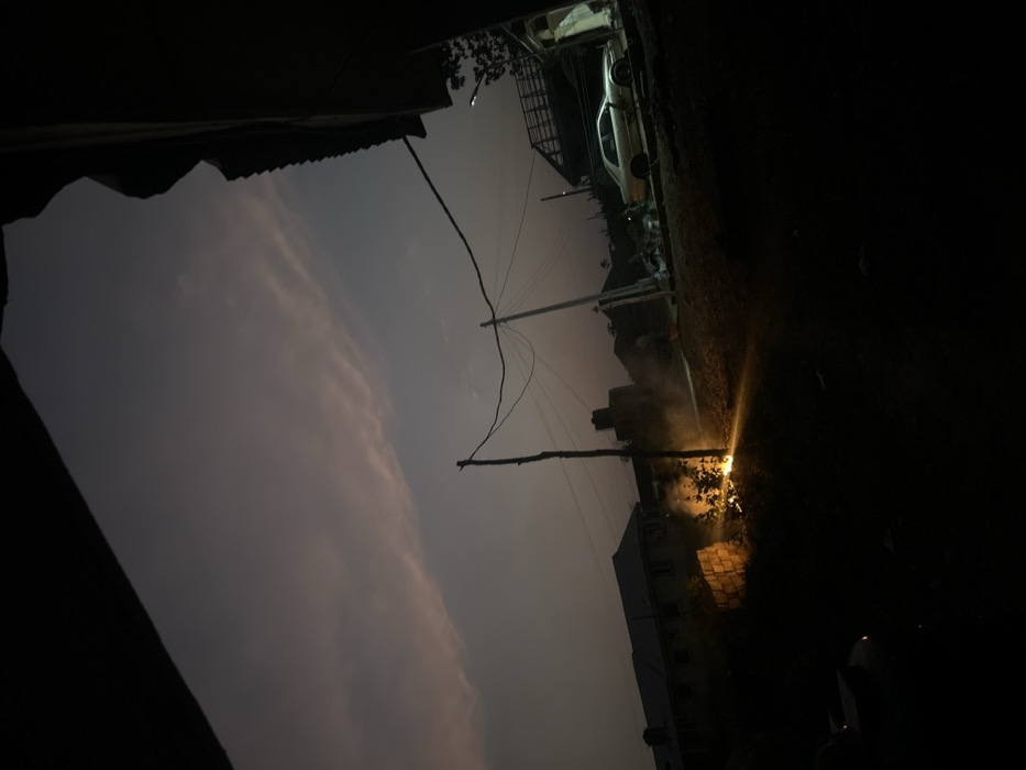 An upward angled view shows a night scene with a predominantly dark sky, partly obscured by a cloud formation. Two wooden utility poles with connecting wires stretch across the frame. On the right, a bright, concentrated light source illuminates a patch of shrubbery, creating visible smoke. Buildings are faintly visible in the background, along with a parked car on top of one. The darkness around these features dominates the composition, suggesting a remote or isolated location.