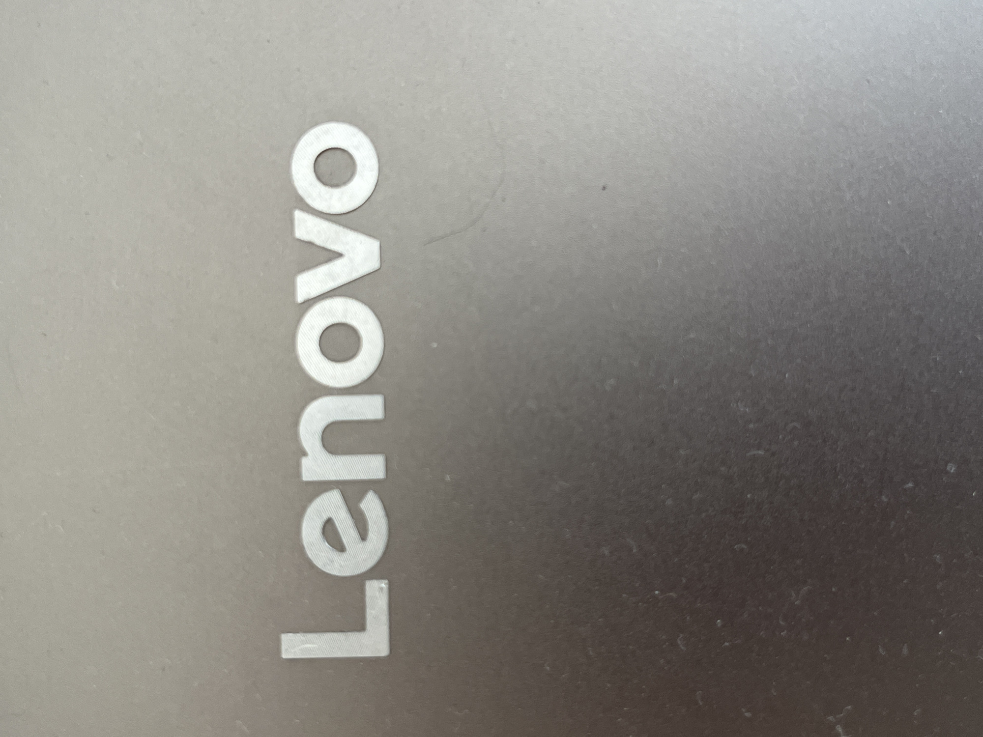 A close-up, high-angle shot shows the "Lenovo" logo on a metallic surface. The letters are white and raised, with a slight sheen. The background is a muted gray with subtle texture and a few dust specks. The lighting is even, creating minimal shadows.