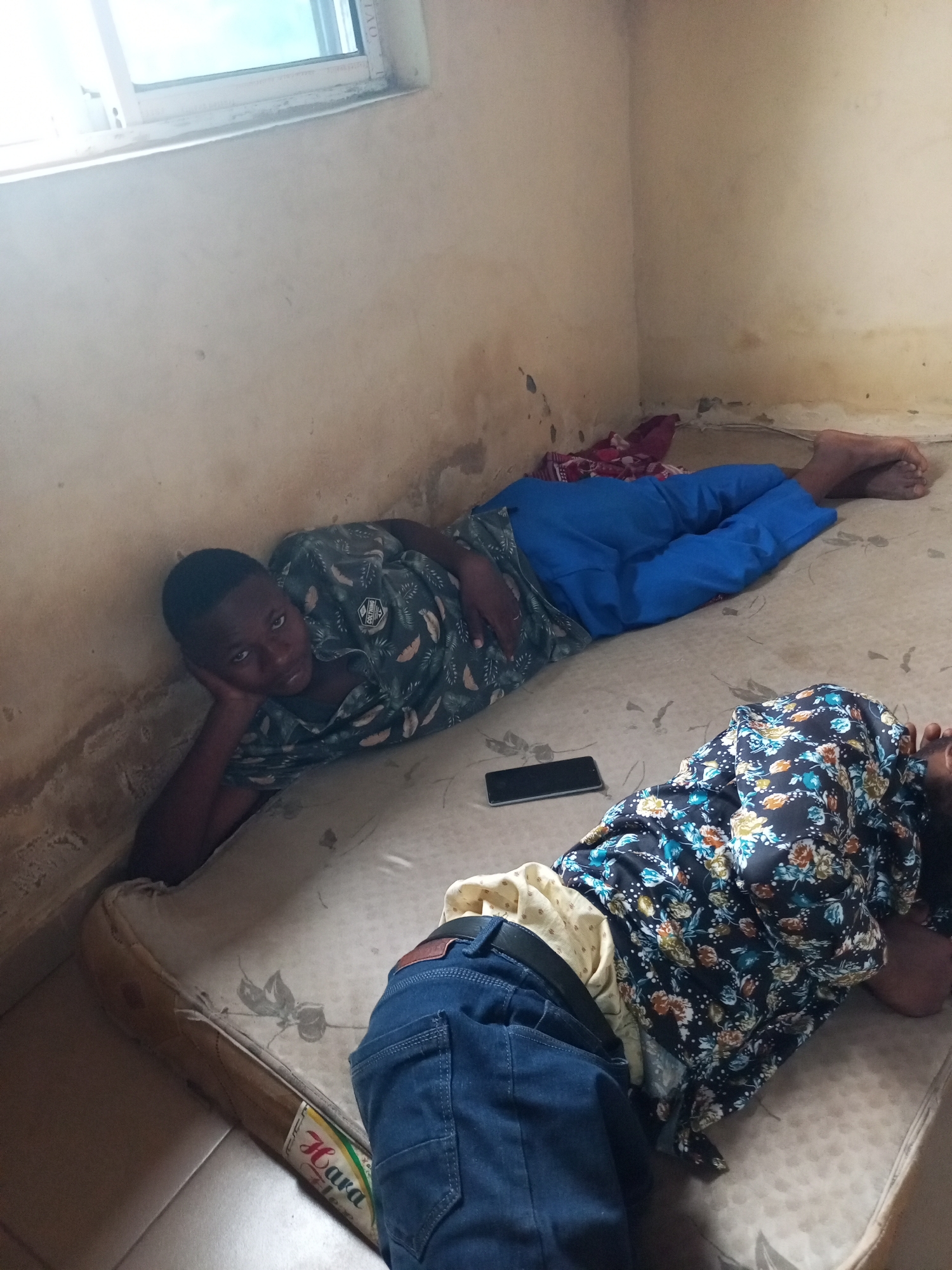 In a dimly lit room, two individuals are lying on a worn mattress placed on the floor. The room appears to be in a state of disrepair, with stained walls and a window overlooking a bright sky. The mattress is covered with a faded floral pattern.
One individual, appearing to be a young male, is lying on his left side, propped up on his elbow, looking towards the camera. He is wearing a dark short-sleeved shirt with a tropical leaf and feather print and blue pants. A mobile phone is placed on the mattress near him.
The second individual is lying on their right side, facing away from the camera, their head resting on the mattress. They are wearing a dark floral-patterned garment and blue jeans.
The scene suggests a simple and possibly impoverished living environment. The presence of the mattress on the floor, the condition of the walls, and the general lack of furnishings point towards basic accommodations.