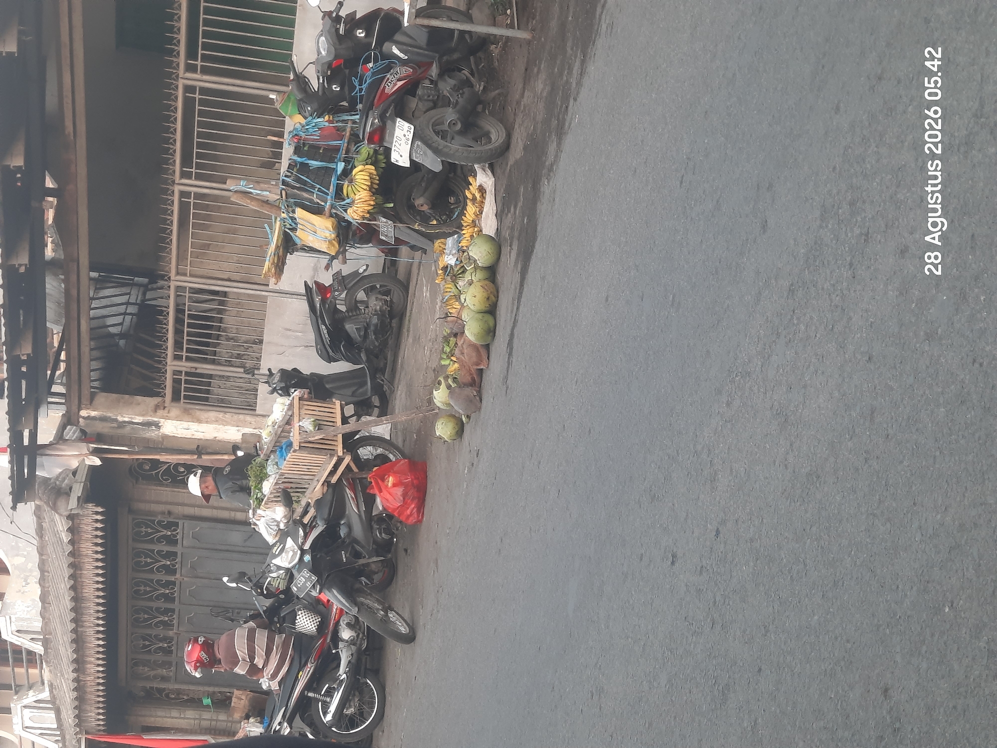 The image depicts a street scene in Pajarakan Kulon, Indonesia, at 05:42 on August 28, 2026. In the foreground, a gray asphalt road occupies the majority of the frame. To the right and center, several motorcycles are parked along the side of the road, some with baskets or racks attached. Produce, including coconuts and bananas, is displayed on the ground and on the baskets, suggesting a roadside market.
Two individuals are visible. One person, wearing a helmet and a striped shirt, is seated on a motorcycle on the left side of the frame. Another person, wearing a white cap and a dark jacket, is standing near a motorcycle with a basket filled with vegetables. The background shows a building with a corrugated roof and metal fencing, typical of urban or semi-urban Indonesian architecture. A red plastic bag is placed on the ground near the motorcycles. The lighting suggests it is early morning.