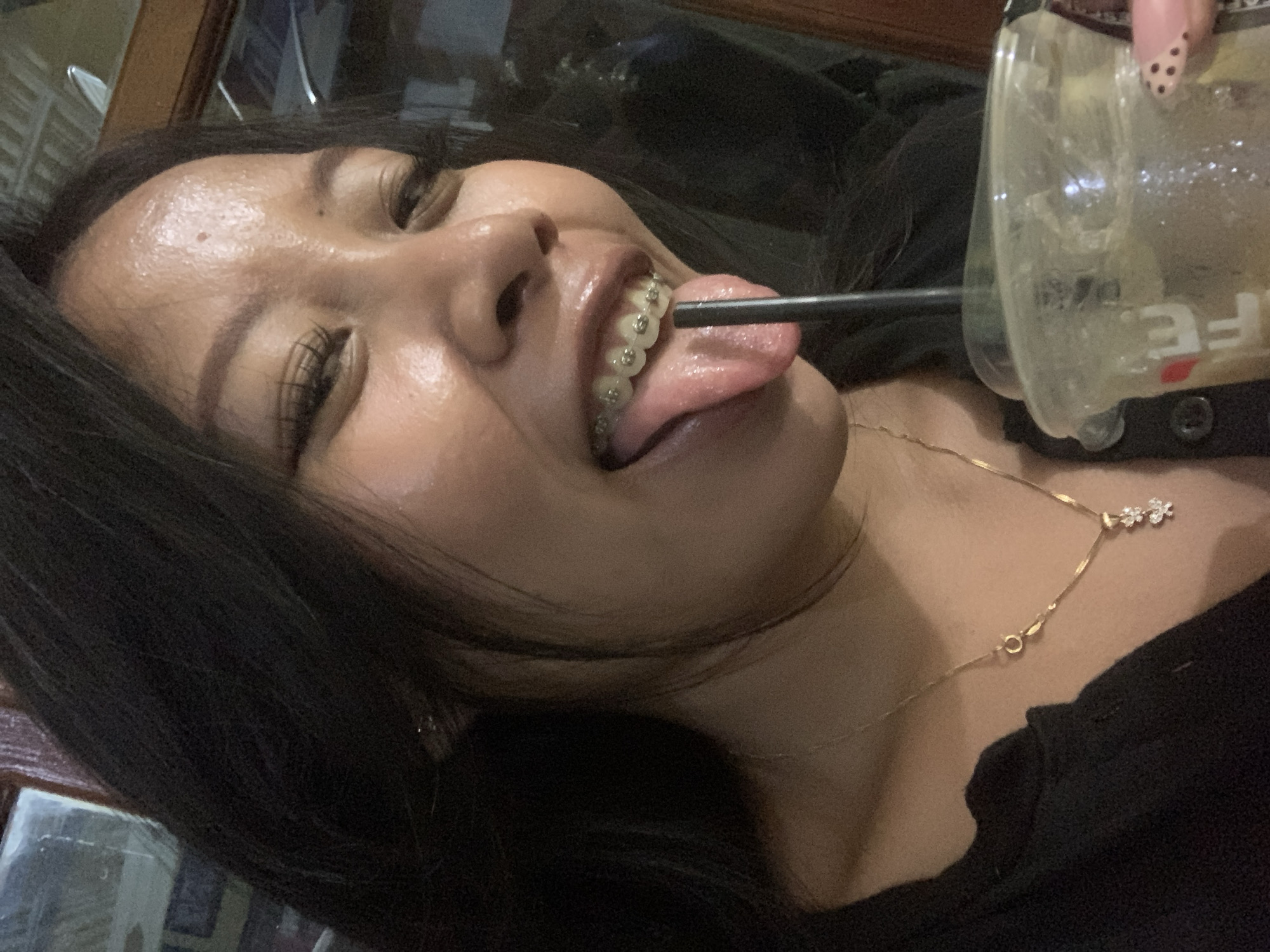 A medium shot captures a young woman with dark hair and braces, sticking her tongue out with a straw in her mouth, from a clear plastic cup. She wears a black shirt and a gold necklace with a small pendant. The background is blurred, but appears to be an indoor setting, possibly a cafe, with wooden elements. The lighting is dim, suggesting it might be evening. There are no visible indicators of the specific location within Jajag, Indonesia.