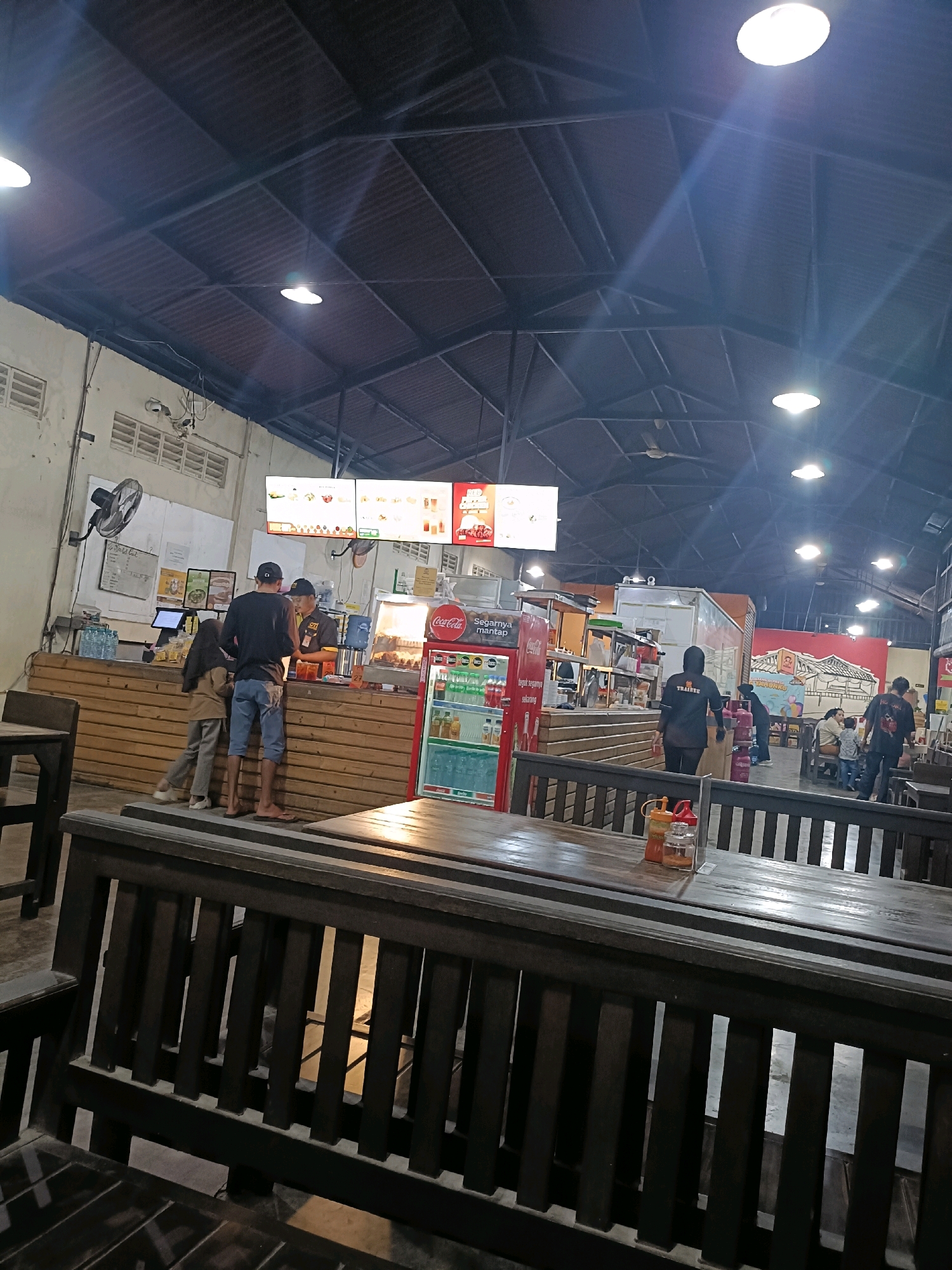 The image depicts the interior of a food establishment, possibly a food court or a casual dining restaurant, located in Semarang, Indonesia. The scene is illuminated by overhead lights, casting a warm glow on the corrugated metal ceiling and the dark wooden furniture. In the foreground, several dark wooden benches and tables are arranged, suggesting a communal dining area.
Further into the establishment, a counter area is visible, where staff members and customers are interacting. Two individuals, a man and a woman, stand at the counter, seemingly placing or receiving an order. A male employee, wearing a dark shirt with a logo and a cap, is attending to them. Behind the counter, a refrigerated display case, branded with "Coca-Cola," contains various beverages. The counter itself is made of light-colored wood. Above the counter, digital menu boards display food items and prices.
To the right, a red refrigerator is prominently placed. Further back, a food truck or a kitchen area is visible, with additional staff members and customers. One staff member, wearing a black shirt with "TRAINEE" printed on it, is walking away from the counter. Another area features a colorful mural on the wall, depicting a traditional Indonesian house and people, adding a local touch to the environment. The overall atmosphere appears to be bustling and casual, characteristic of a popular eatery.