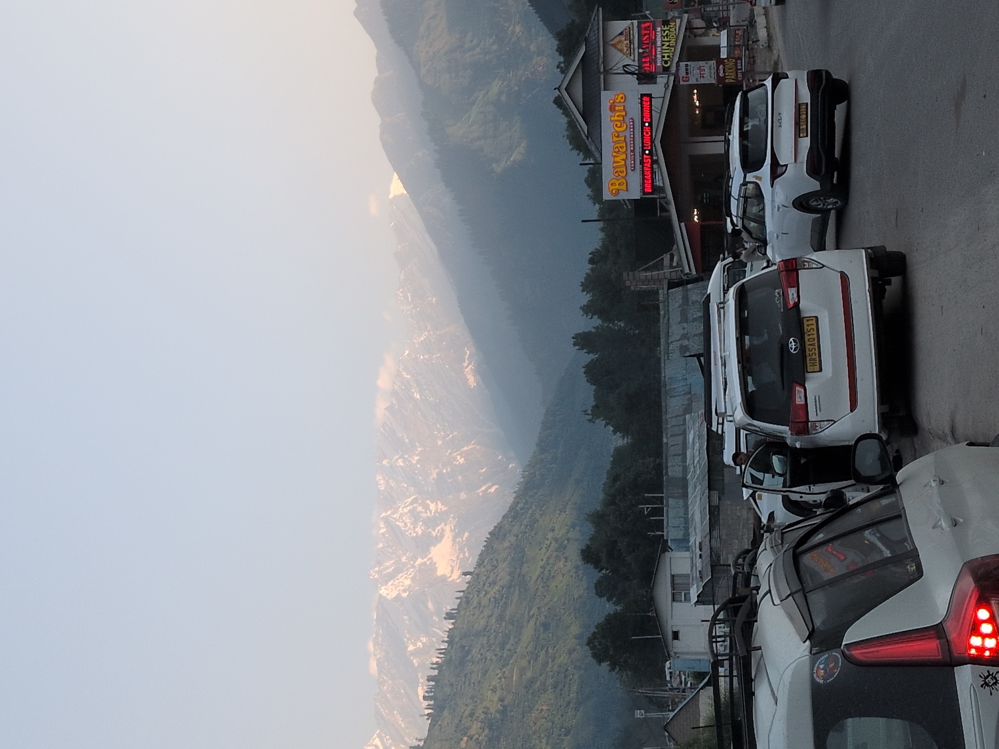 The image depicts a street scene in Vashisht, India, with mountains in the background. In the foreground, several white vehicles are parked or moving on a road. One vehicle has its driver's side door open, and a person is visible inside. Another vehicle has a license plate reading "HR55AQ1511". A prominent sign for "Bawarchi's Family Restaurant" is visible on a building, with text indicating "BREAKFAST LUNCH DINNER". Other smaller signs for "Cafe Dho" and "NORTH INDIAN CHINESE INDIAN" are also present. The environment suggests a hilly region with green slopes leading up to snow-capped mountains in the distance, under a hazy sky.