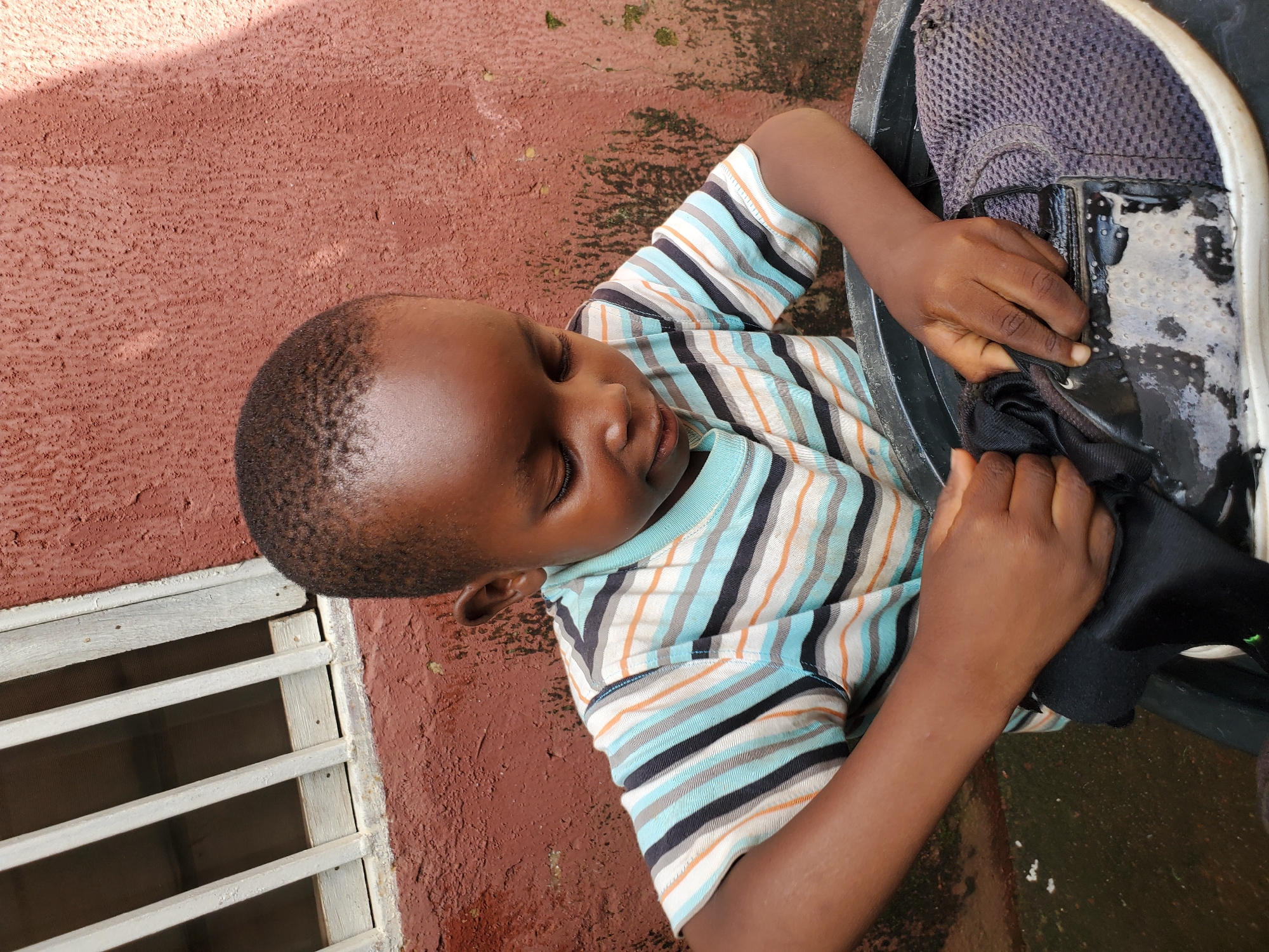 A young boy, with closely cropped dark hair and dark skin, is shown from a high angle, looking down with his eyes closed and head bowed. He is wearing a striped t-shirt with horizontal bands of light blue, orange, black, and white. His hands are engaged with a dark-colored shoe submerged in a black basin. The shoe appears to be a sneaker with a mesh texture on the side and a black material on the front, possibly leather. The basin is filled with soapy water, evident from the suds and reflections on the shoe's surface.
The background consists of a rough, textured reddish-brown wall, which appears to be an exterior surface. To the left, a section of a window with white bars and a screen is visible, set against the same wall. The lighting suggests it is daytime, with some shadows cast on the wall. The overall scene is consistent with an outdoor setting in a residential or common area within the city of Jos, Nigeria. The damp ground or surface below the basin is visible in the lower portion of the image, with scattered white specks that might be soap residue or debris. The boy's posture and expression suggest he is concentrating on cleaning the shoe.