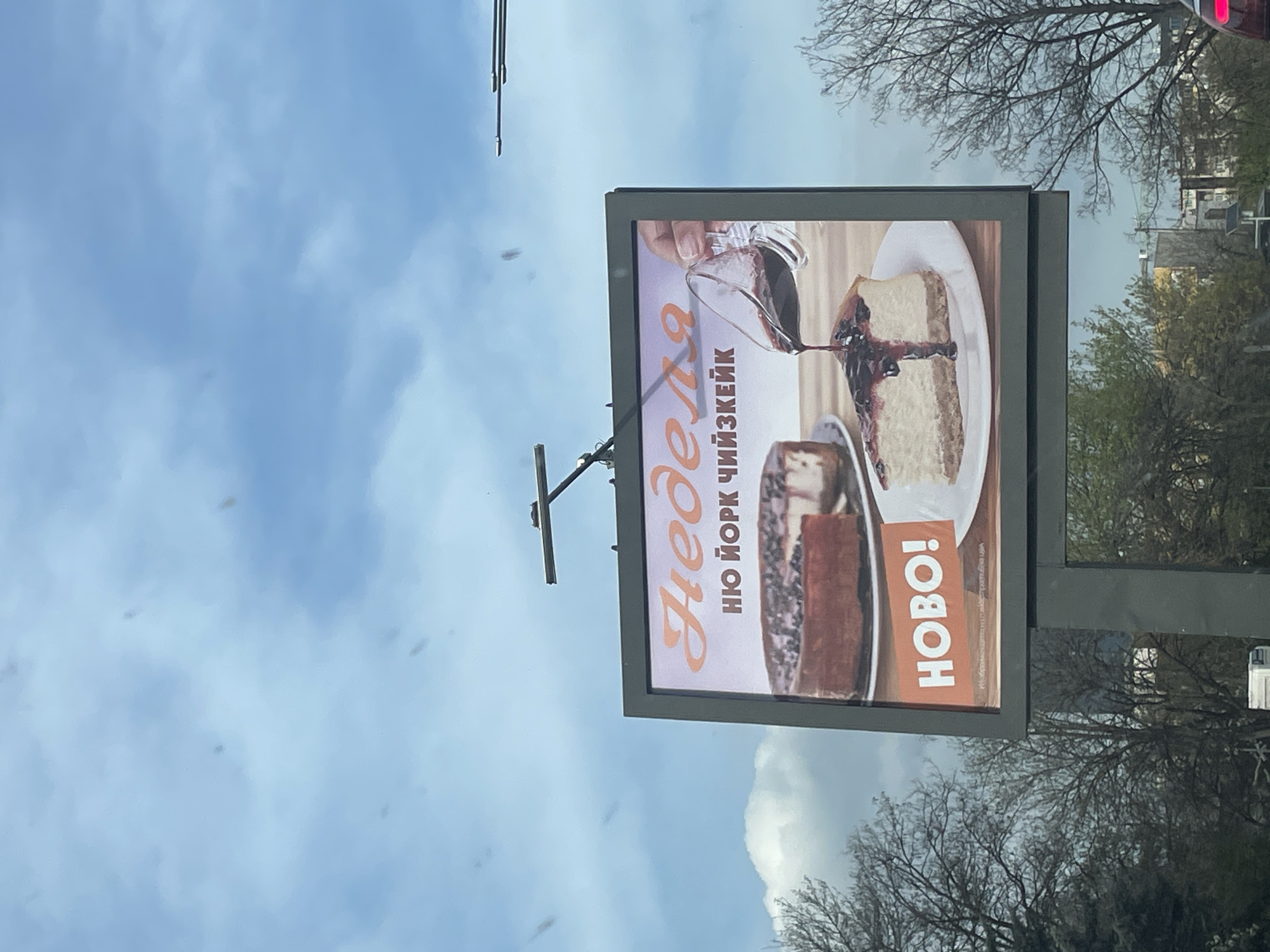 An outdoor scene captures a large billboard against a cloudy daytime sky. The image appears to be taken from a vehicle, with some visual artifacts resembling specks of dust or water on a window. The billboard, oriented vertically, prominently displays an advertisement for cheesecake.
On the billboard, a close-up image shows a hand pouring a dark sauce, possibly fruit topping or chocolate, over a slice of New York-style cheesecake placed on a white plate. Below it, another slice of chocolate cake is partially visible. The background of the advertisement features a warm, light brown tone.
Visible text on the billboard is in Cyrillic. The main brand name reads "Неделя" (Nedelya), which is a known patisserie chain. Below this, "НЮ ЙОРК ЧИЙЗКЕЙК" (New York Cheesecake) describes the product, and "НОВО!" (Novo!), meaning "New!", highlights it as a new offering.
The setting behind the billboard includes bare-branched trees, typical of late winter or early spring, with hints of some new green foliage appearing. In the distance, through the branches, parts of buildings or structures can be vaguely seen. The sky is largely overcast with soft, light blue clouds. The overall impression is an urban or roadside environment in Sofia, Bulgaria, as indicated by the location context.