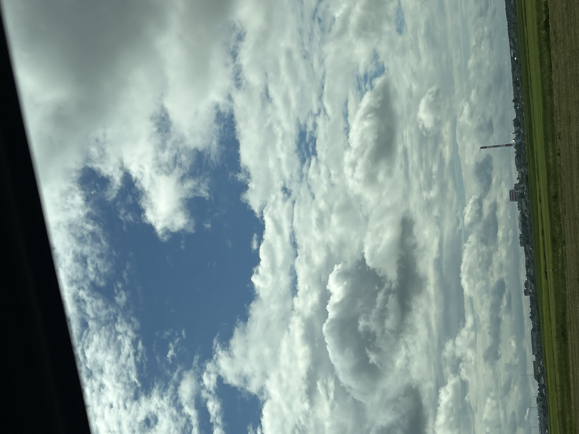 The image depicts a wide view of the sky over a rural landscape with a town or village in the distance. The sky is filled with dramatic, fluffy white and grey clouds against a backdrop of a bright blue sky. In the lower portion of the image, a strip of green fields is visible, suggesting a natural or agricultural setting. Beyond the fields, a cluster of buildings forms a settlement, with a prominent red and white striped smokestack or industrial tower standing out against the buildings and the sky. The overall impression is of a daytime scene with varied cloud cover. The location is specified as Урыцкі сельскі Савет, Belarus.