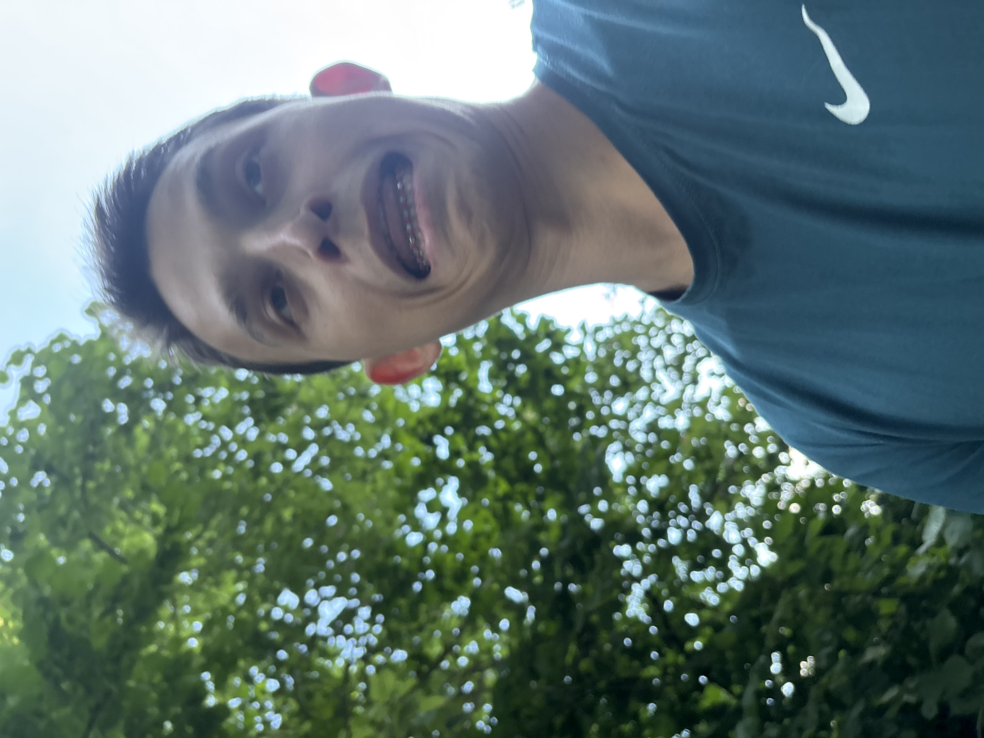 In this selfie, a young person with a wide, playful grin is captured from a low angle, looking up at the camera. Their mouth is open, revealing teeth and orthodontic braces, adding a touch of youthful individuality to the image. The person is wearing a dark teal t-shirt with a white Nike logo visible on the right side. The background is a blur of vibrant green foliage and bright white sky, suggesting an outdoor setting on a sunny day. The greenery, possibly from trees, is out of focus, creating a bokeh effect that draws attention to the subject's face. The lighting indicates it is daytime, likely late morning or early afternoon, given the brightness of the sky. The overall mood is lighthearted and energetic, conveyed through the person's exaggerated facial expression. The scene is set outdoors, possibly in a park or garden, with the sun filtering through the leaves, creating dappled light. There are no visible text elements other than the Nike logo on the shirt.