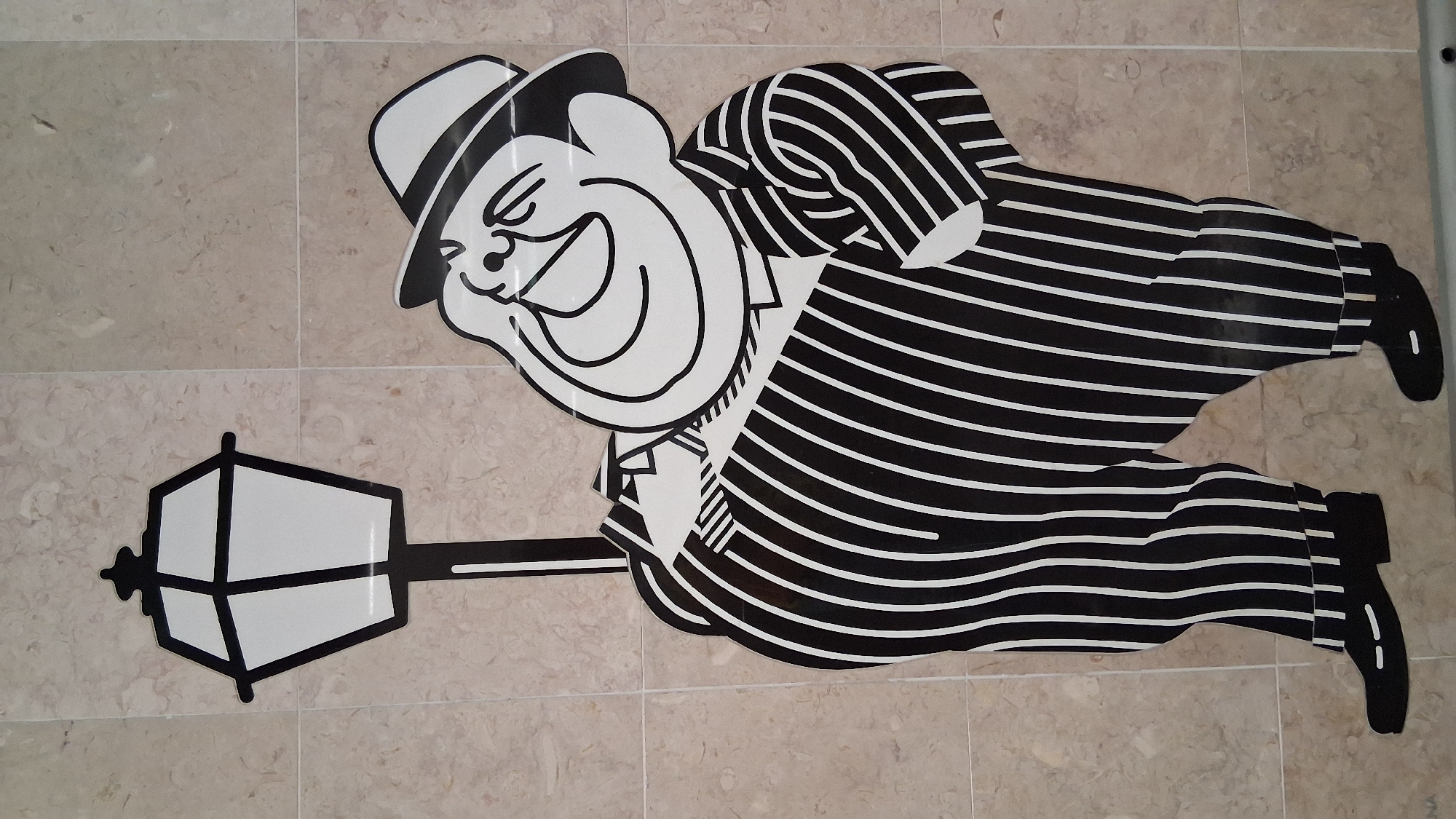 A cartoon depiction of a corpulent man in a pinstripe suit and fedora stands next to a lamppost. The man is laughing with his head tilted back. The artwork is adhered to a tiled wall. The bottom right corner of the image shows a faint inscription that reads "LUZ". The scene is likely located in Lisbon, Portugal, as the word "LUZ" translates to "light" in Portuguese and is also the name of a famous square and metro station in Lisbon.