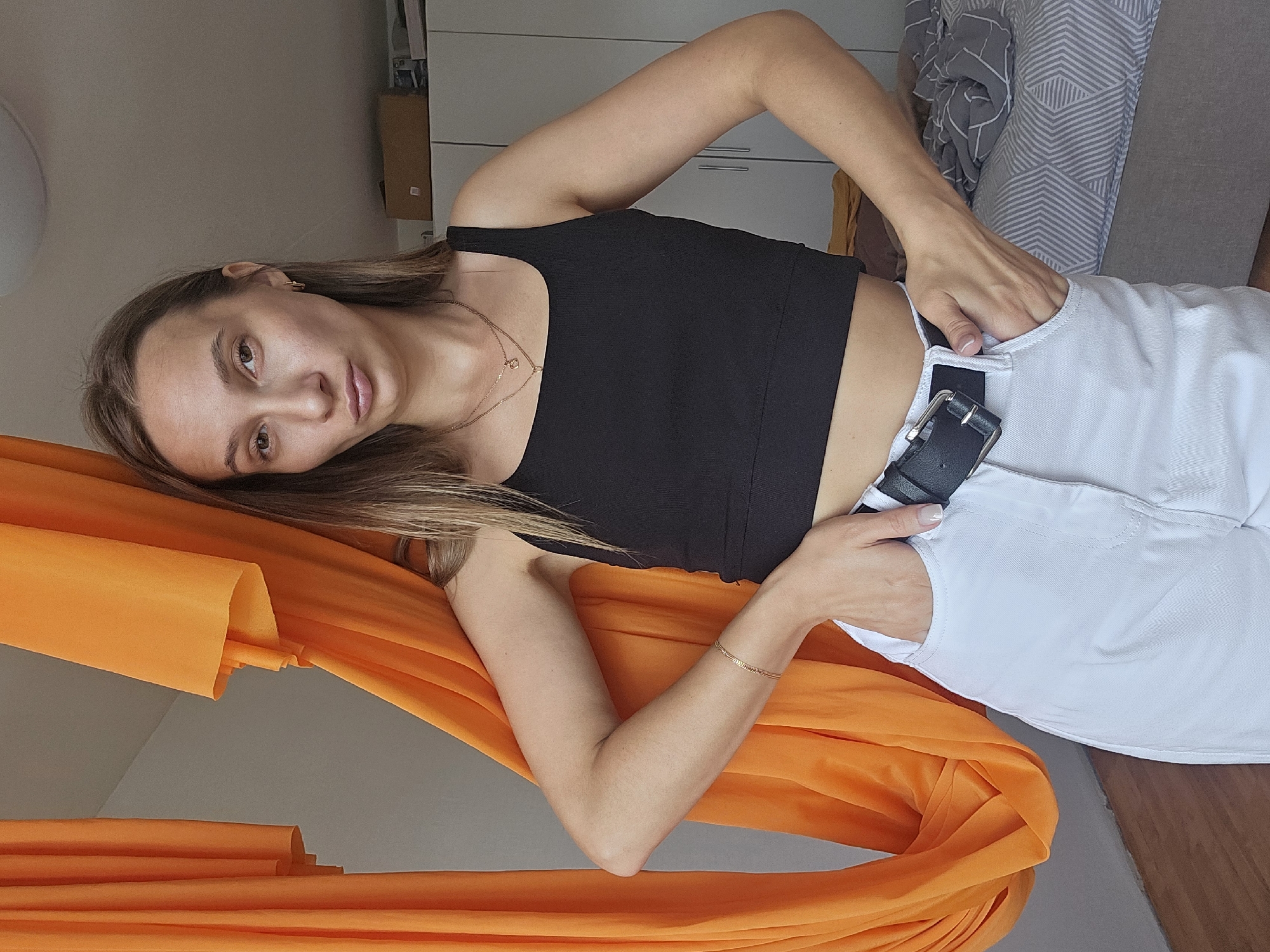 A young woman with long brown hair and light skin stands in an indoor setting, possibly a bedroom. She is wearing a black crop top, white high-waisted pants, and a black belt with a silver buckle. She has her hands in her pockets and is looking at the camera with a slightly puckered expression. To her left is a large piece of bright orange fabric, draped and folded. In the background, a bed with gray and white striped bedding is visible, along with a closet with white doors. The lighting is natural and appears to be coming from a window or overhead source. There is no indication that this scene is located in Chaikovsky, Russia.