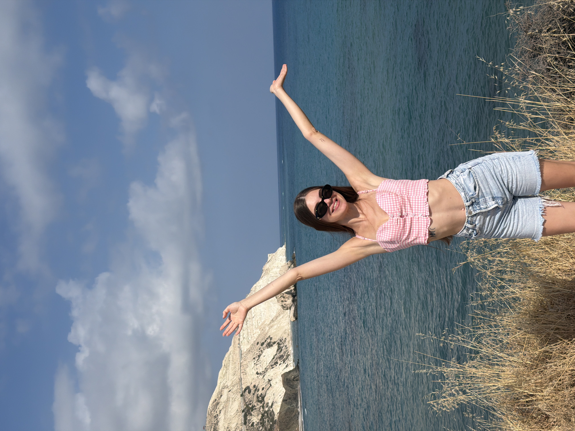 This is a medium shot of a young woman with long brown hair, wearing sunglasses, a pink and white checkered crop top, and distressed denim shorts. She is standing on a cliff overlooking the ocean, with her arms outstretched and raised in a joyful gesture. The sea is a deep blue, and the sky is clear with scattered white clouds. Dry, golden grass lines the edge of the cliff in the foreground, adding texture to the scene. The woman's expression is one of happiness and freedom, amplified by her pose and the expansive natural backdrop. The lighting suggests it is daytime, likely sunny, with the sun casting a soft light on the scene. The terrain of the cliff face is rugged and rocky. The overall atmosphere is one of exhilaration and enjoyment of a beautiful outdoor setting.