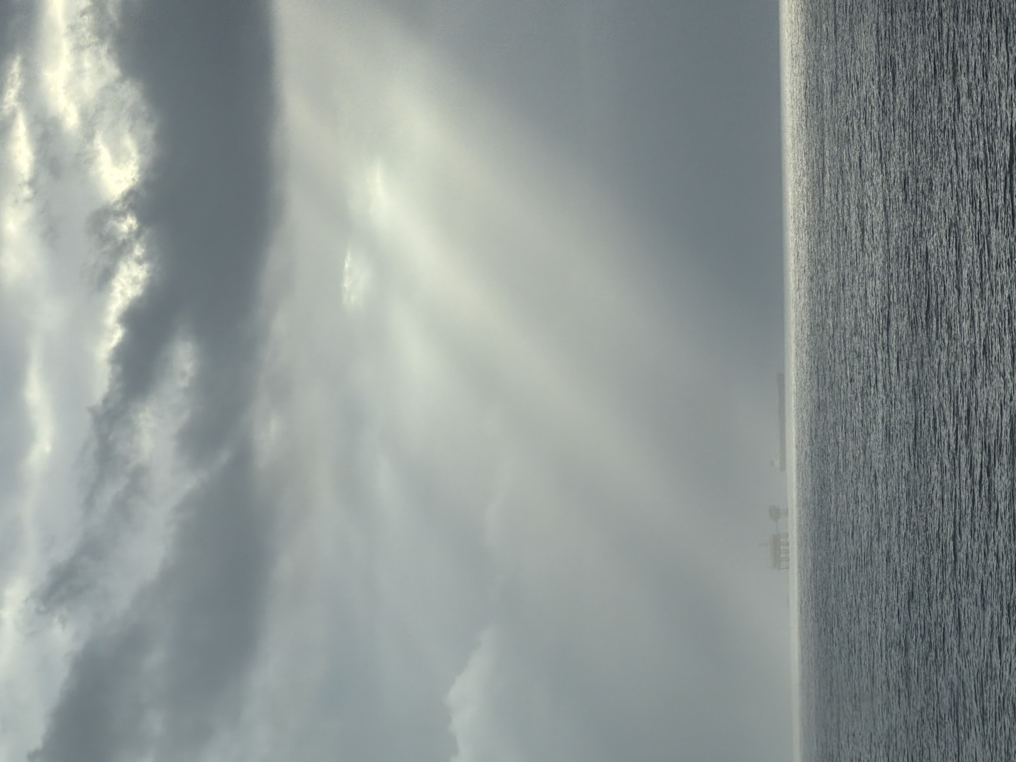 The image, oriented vertically, depicts a dramatic seascape with a striking sky and expansive water. When viewed with the horizon set horizontally, the upper half reveals a dynamic sky filled with thick, grey clouds. Brilliant shafts of sunlight, known as crepuscular rays or "god rays," pierce through the cloud cover, creating vivid streaks of light that highlight areas against the darker cloud formations.
The horizon line is clearly defined, separating the sky from a vast body of water. The lower half of the image displays this water, appearing in muted grey-blue tones. Its surface is highly textured with fine, parallel lines, suggesting ripples, small waves, or currents on a large body of water.
In the far distance, faintly visible on the horizon line just above the water, are several indistinct structures. These appear to be man-made, possibly resembling offshore platforms, industrial rigs, or a distant pier, their details obscured by distance and atmospheric conditions.
The scene captures a moment of changing weather over a large sea or ocean. The prominent sunbeams breaking through clouds suggest either sunrise, sunset, or a period when the sun emerges after heavy cloudiness. No people or specific activities are directly visible. There is no discernible text within the image. The overall atmosphere evokes a sense of natural grandeur and tranquility.