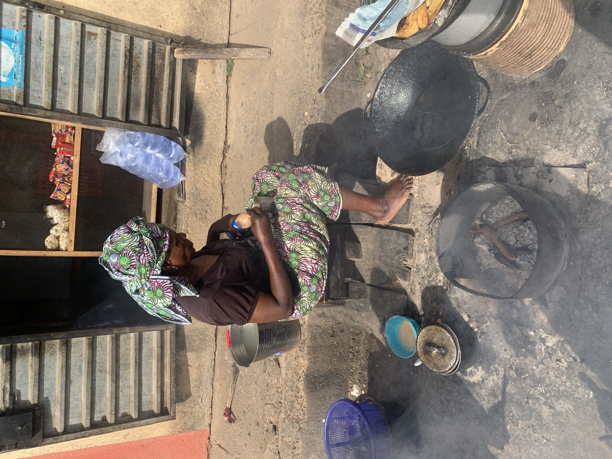 An outdoor scene in Jos, Nigeria, features a woman engaged in cooking and possibly selling food by a roadside or stall. The image is taken from a high angle, looking down.
The primary subject is a woman, likely of middle age, seated on a low wooden stool. She wears a dark brown top and a vibrantly patterned skirt, complemented by a matching headscarf with green, white, and maroon motifs. Her bare feet are visible, indicating an informal setting. She is focused on an activity with her hands, possibly preparing ingredients or tending to a small item she holds.
The cooking setup includes large, dark cast-iron pots or woks, one of which is empty and another possibly containing food (some yellow items, possibly fried plantains or potatoes, are visible in a pot lined with newspaper in one of the crop hints). An open fire pit, made from a cut-off barrel or drum, burns wood, emitting smoke. Several other items are scattered around: a light blue plastic bowl with some liquid, a smaller lidded pot, a blue plastic mesh basket, and a metal bucket filled with water. A small, crude broom lies on the ground.
To the left, a stall or shop structure with a corrugated metal facade is partially visible. Inside, through a mesh opening, packets of snacks or instant noodles and small white items (possibly popcorn or another snack) are displayed for sale. Bags of bottled sachet water, clearly labeled "SACHET WATER," are stacked near the opening. Above these, a blue package with text like "Everyday Clay" and "35g" is also visible.
The ground is unpaved, a mix of dirt and rough concrete, showing signs of wear and activity. The strong sunlight and pronounced shadows suggest it is daytime, likely morning or afternoon, with clear weather. The overall ambiance points to a bustling, everyday street or market environment where food is prepared and sold.