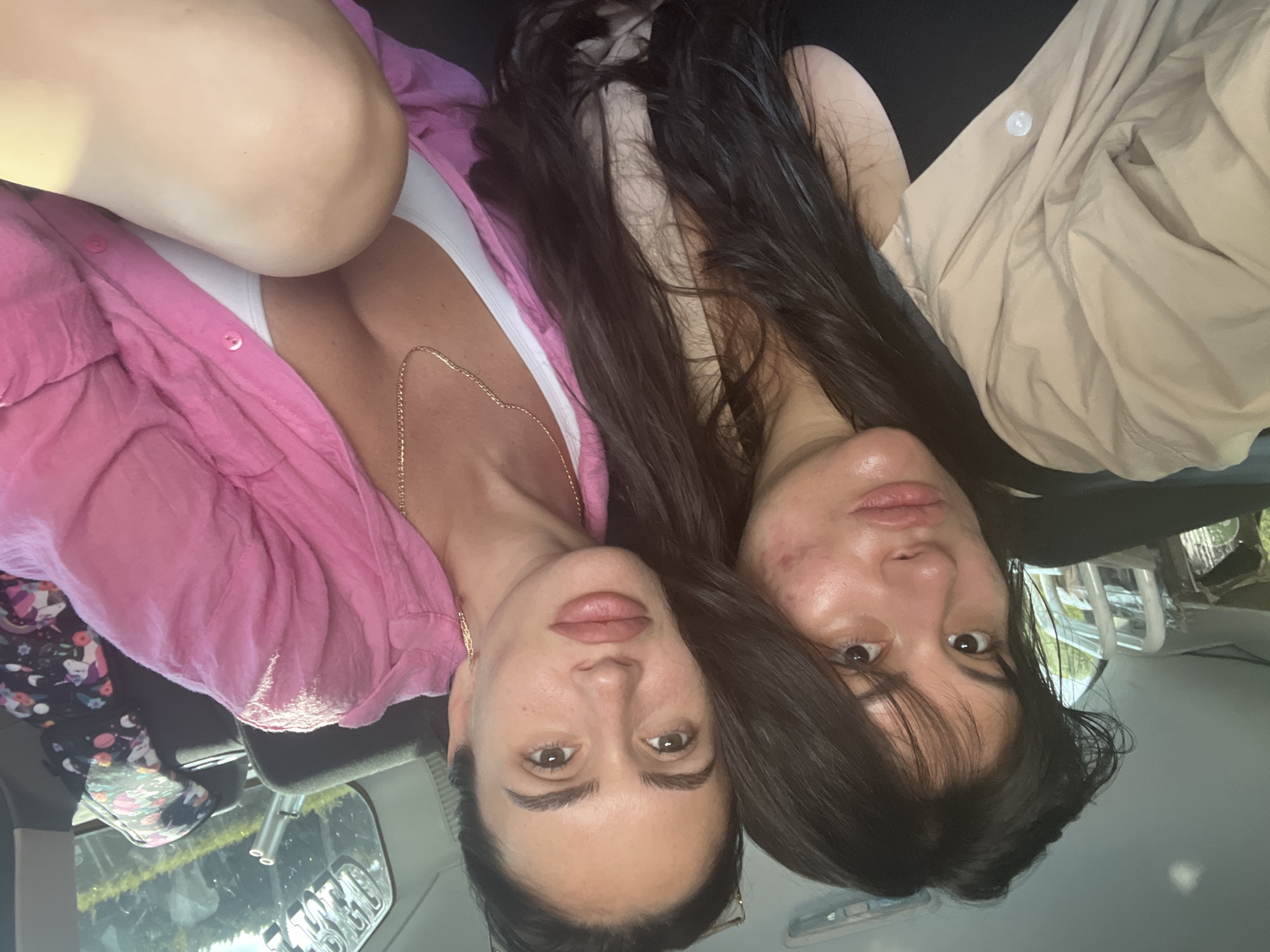 Two women are taking a selfie inside a vehicle. The woman on the left has dark hair and is wearing a tan shirt over a black top. The woman on the right has dark hair and is wearing a pink shirt over a white top, with a gold necklace. The background shows the interior of a car and a blurred view of trees and sky through the windows. The image appears to have been taken outdoors on a sunny day.