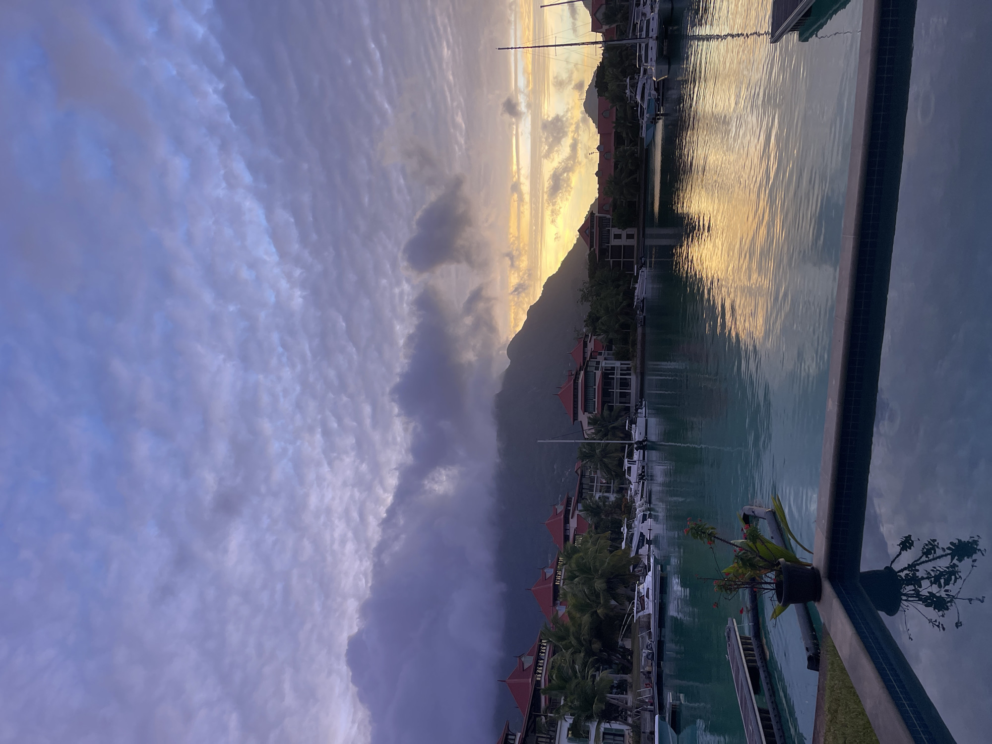 This image depicts a serene waterfront scene, likely at dawn or dusk, at Eden Island Marina in Victoria, Seychelles. The foreground features a swimming pool with tiled edges, its water reflecting the dusky sky and a potted plant. Beyond the pool, a body of calm, turquoise water serves as a marina, dotted with several moored boats, including yachts and speedboats. Along the far bank, a row of buildings with distinctive red-tiled roofs and white facades lines the waterfront, interspersed with lush palm trees. In the background, a majestic mountain range, partially obscured by dramatic, cloud-laden skies, adds a sense of depth and grandeur to the landscape. The sky is a canvas of soft purples, blues, and a streak of warm yellow where the sun is either rising or setting, creating a tranquil and picturesque atmosphere. The overall impression is one of a luxurious and peaceful tropical resort.