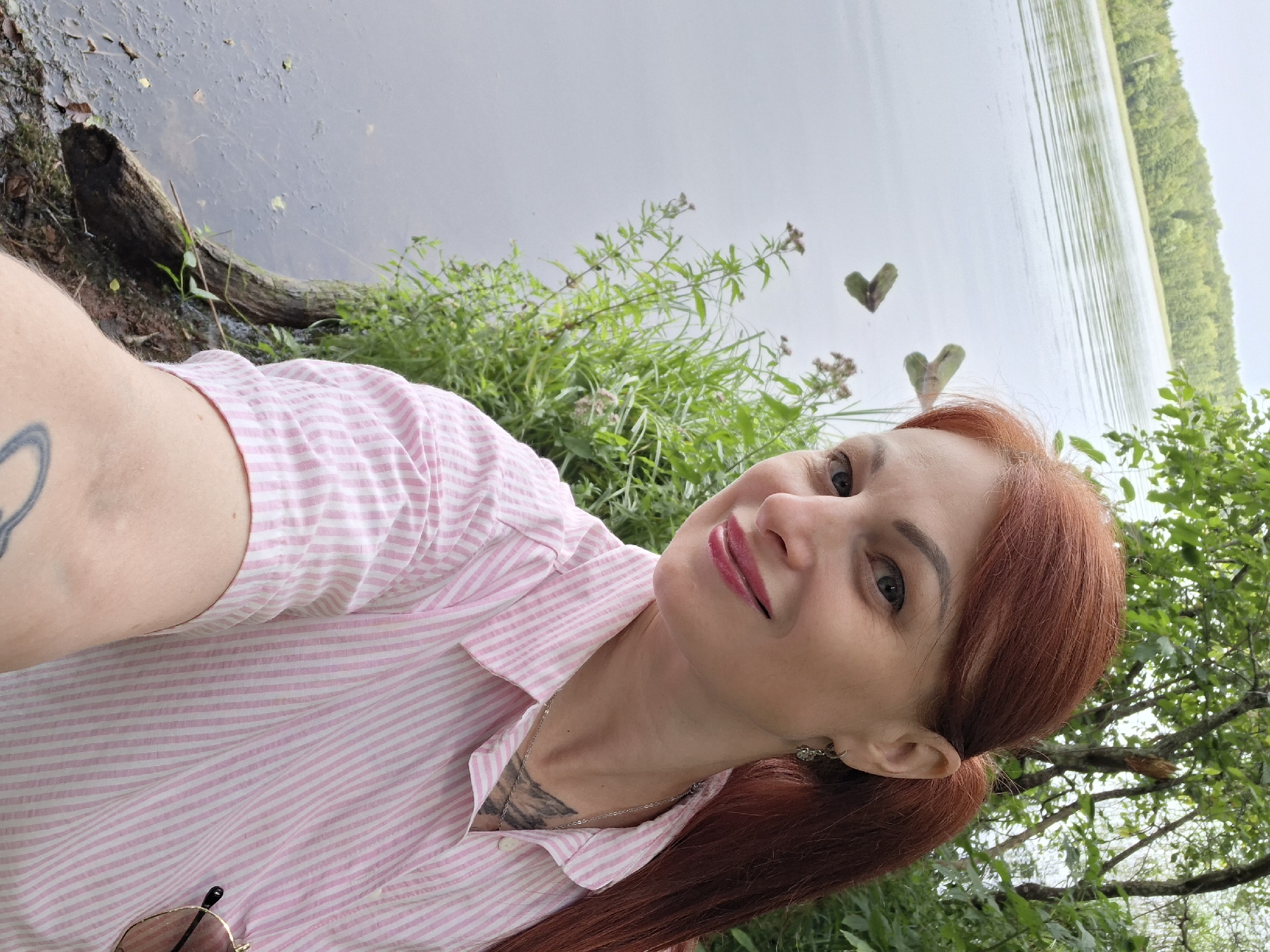 In a selfie taken by a woman with red hair, she is positioned on the right side of the frame, looking at the camera with a smile. She has makeup on, including lipstick. She is wearing a pink and white striped shirt and a necklace. A tattoo is visible on her chest and arm. To her left is a body of water, possibly a lake, with greenery on the far bank. The immediate foreground shows the edge of the water with some wood debris and vegetation. The scene appears to be outdoors, likely in a natural setting such as a park or lakefront. The location is identified as Мядзельскі сельскі Савет, Belarus.