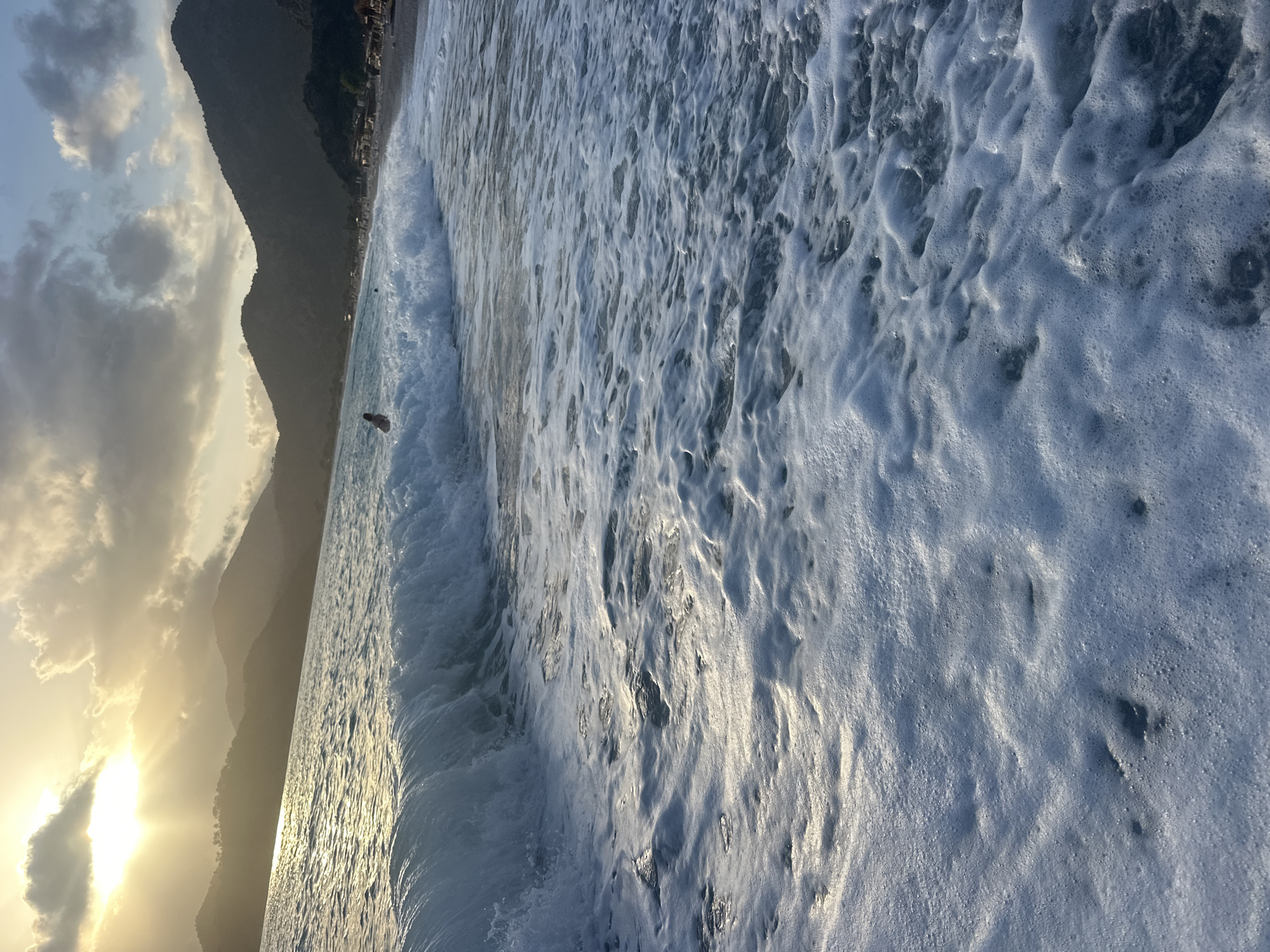 The image presents a beach scene, likely in Fethiye, Turkey, given the presence of coastal mountains and the city stretching along the shoreline. The immediate foreground is dominated by a breaking wave, with white foam covering a large portion of the frame. The water has a bluish-grey tone.
Further into the frame, a person is seen standing in the water. In the background, mountains stretch along the coast, with buildings indicating an urban presence. The sky is partially cloudy, with the sun low on the horizon, casting a golden light.