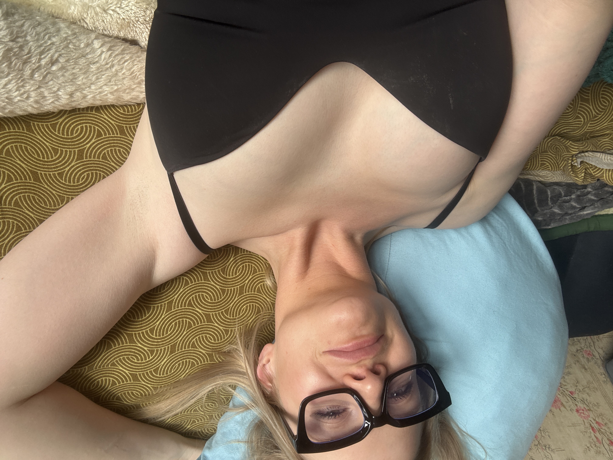 A close-up, high-angle shot captures a woman lying down indoors in Saint Petersburg, Russia. Her head is positioned at the bottom of the frame, facing upwards towards the camera, while her body extends towards the top. She has blonde hair and fair skin, and is wearing large black-framed glasses, through which her eyes are visible. Her expression is subtle, conveying a serene or relaxed mood with a hint of a smile. She is dressed in a black top or bra with a distinct, angular neckline.
Her head rests on a light blue pillow. To her left side, an olive green fabric with a repeating golden swirling pattern covers part of the surface she is lying on. In the top left corner, a textured, light beige, fluffy material is visible. Other fabrics, including a floral patterned one and a dark solid one, can be seen in the background, suggesting a cozy, domestic setting like a bedroom or living area. No text is visible in the image. The scene suggests either a moment of rest or a posed portrait.