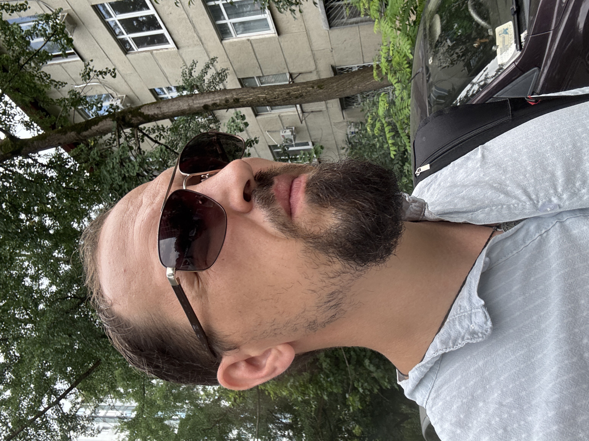 The media file shows a close-up, vertically oriented, outdoor shot of a person's face and upper body. The person is wearing sunglasses with dark lenses and a light blue, button-down shirt. The background is blurred, featuring greenery from trees and the facade of a light-colored building with multiple windows. There are also hints of a dark-colored vehicle. The lighting suggests it is daytime. The person has short, dark hair and a beard. The scene appears to be set in an urban environment, possibly a residential area, given the building and trees. The image's orientation is rotated, making it appear sideways. The overall mood is casual and candid. There is a partial view of a license plate on the vehicle, but no legible text is discernable. No specific activity or event is clearly depicted beyond the subject being outdoors and posing for a photo. The location context of Sochi, Russia, implies a potentially warm climate.