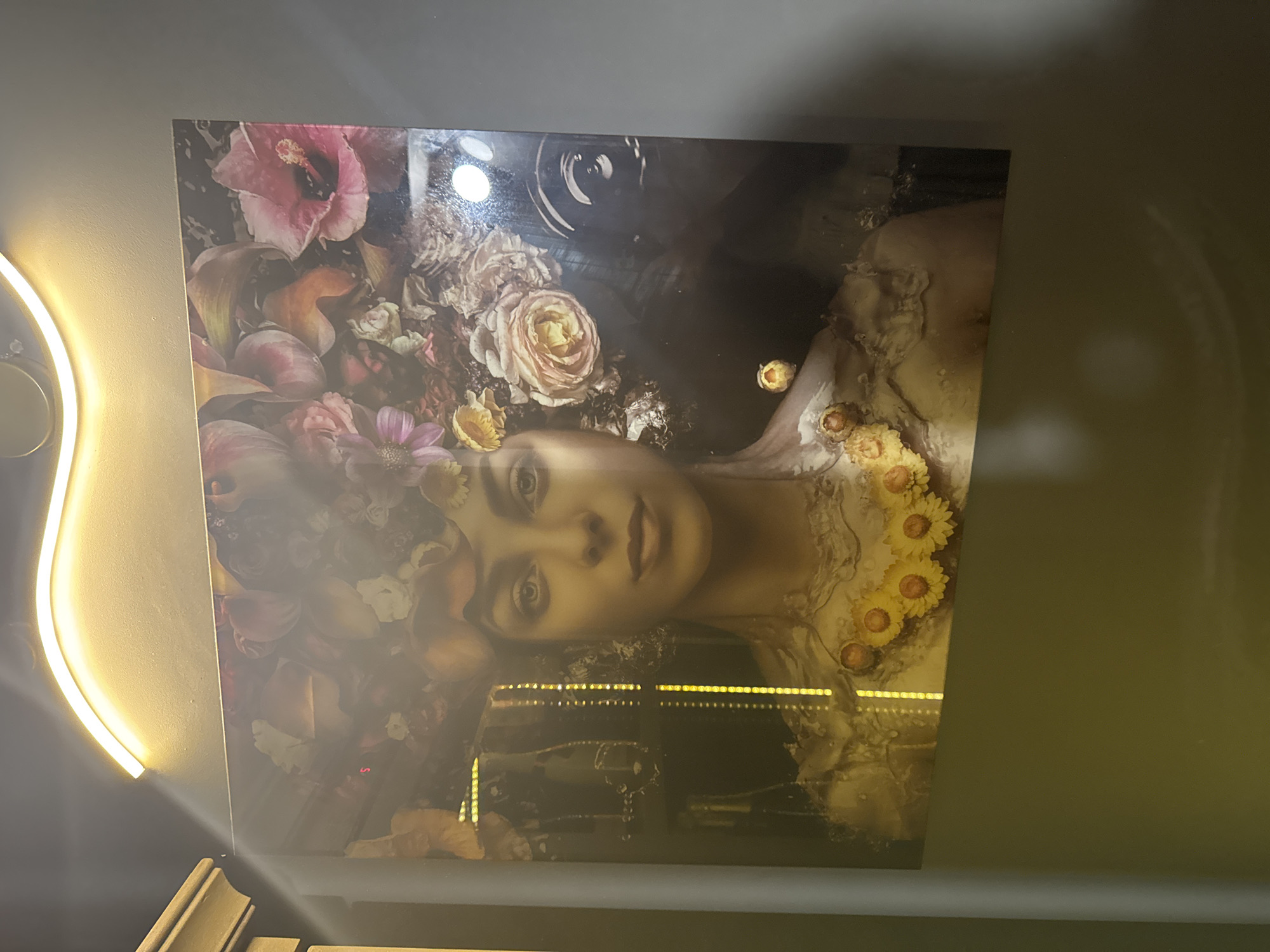 The image displays a framed artwork featuring a woman adorned with flowers. The artwork is positioned on a wall, with a glowing overhead light fixture casting a warm illumination on the scene. The backdrop appears to be a dark, textured wall. The artwork itself depicts a woman with a serene expression, her hair intertwined with a vibrant arrangement of flowers, including pink hibiscus, roses, and smaller yellow blossoms forming a necklace. The artistic style is a blend of realism and surrealism, with a painterly quality. The environment appears to be indoors, possibly a private residence or a gallery. Based on external information, this artwork is located in Sofia, Bulgaria.