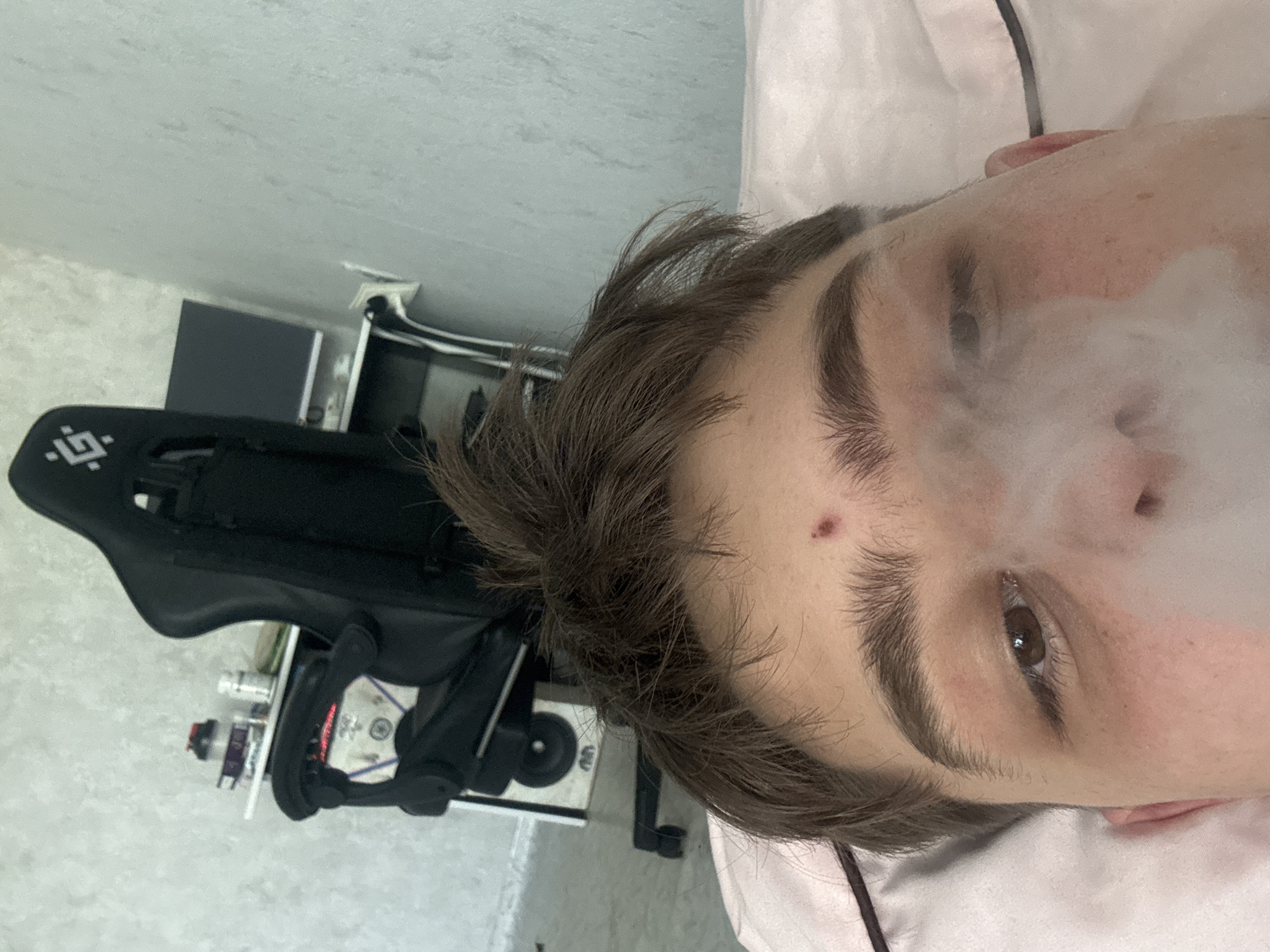 A close-up, slightly rotated perspective depicts a person with brown hair and eyebrows, with smoke obscuring part of their face. A small red blemish is visible on the forehead. Adjacent to the person is a black object with a complex design and what appears to be a white box with several items on top, including a black cable. The background includes a white wall and a white surface, possibly a floor. The location in the city of Unknown, Unknown is not determinable from the image.
