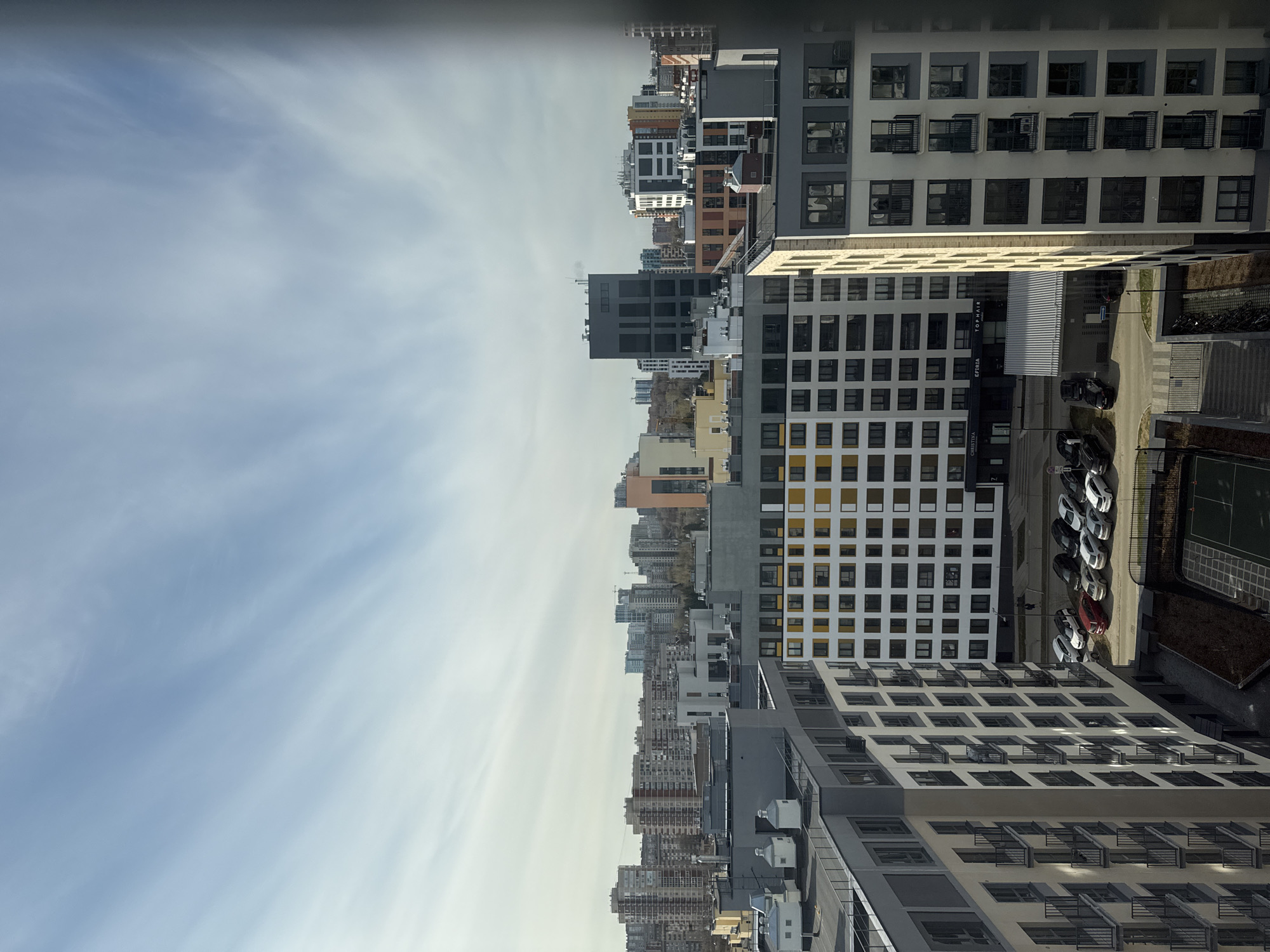 This panoramic image, correctly oriented (rotated 90 degrees counter-clockwise from its vertical presentation), captures a daytime view of a dense urban cityscape in Izhevsk, Russia.
The sky occupies the upper portion, displaying a pale blue hue largely covered by thin, wispy white clouds. The lighting is bright and diffused, suggesting a soft daylight, possibly mid-morning or afternoon, with subtle shadows cast by buildings and vehicles.
The lower two-thirds of the frame are filled with numerous modern, multi-story buildings, primarily appearing to be residential or commercial complexes. These structures exhibit a variety of architectural styles, predominantly featuring clean lines, repeating patterns of square windows, and dark-railed balconies. Building facades are mostly in shades of white, beige, grey, with some sections accented in dark grey, yellow, or orange. The density of buildings suggests a developed urban core or a large residential district.
In the foreground and mid-ground on the right, several distinct buildings are prominent. Below them, a paved parking lot holds a line of parked cars, including white, black, and red vehicles. Adjacent to the parking area is a sports court, identifiable by its green surface and white demarcation lines, possibly for tennis or basketball, enclosed by a fence.
No people are explicitly visible. The scene is static, capturing the architectural landscape of the city.
Visible text on one of the buildings includes "TOP HAIR" and "CHRISTINA," with "CPETA" (or similar, partially obscured) between them. Another small sign displays the text "NIZ."