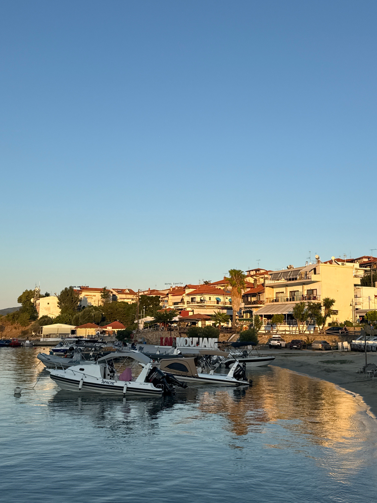 The image shows a waterfront scene in Ammouliani, Greece, during what appears to be late afternoon or early evening. The sky is clear and blue, with the sun casting a warm glow on the water and the buildings.
In the foreground, several boats are moored in the calm, blue water. One white inflatable boat prominently displays the name "APACHE" on its side. Other boats, some with canopies, are visible further back. The water reflects the colors of the sky and the surrounding environment, creating shimmering patterns.
Along the shoreline, a sandy beach is visible, with lounge chairs and tables set up, suggesting a seating area for a restaurant or cafe. The water gently laps the shore. In the background, a cluster of buildings forms the town's skyline. These buildings are primarily white and light-colored with terracotta roofs, typical of Mediterranean architecture. Signs on the buildings indicate businesses such as "HOTEL AMMOULIANI" and "HOTEL SUNRISE." Green trees and vegetation are interspersed with the buildings, particularly on the gentle slope rising behind the town. A large sign with the letters "IAM MOUJAM" is partially visible near the beach. Several cars are parked along the road behind the beach area. The overall impression is one of a peaceful coastal town.