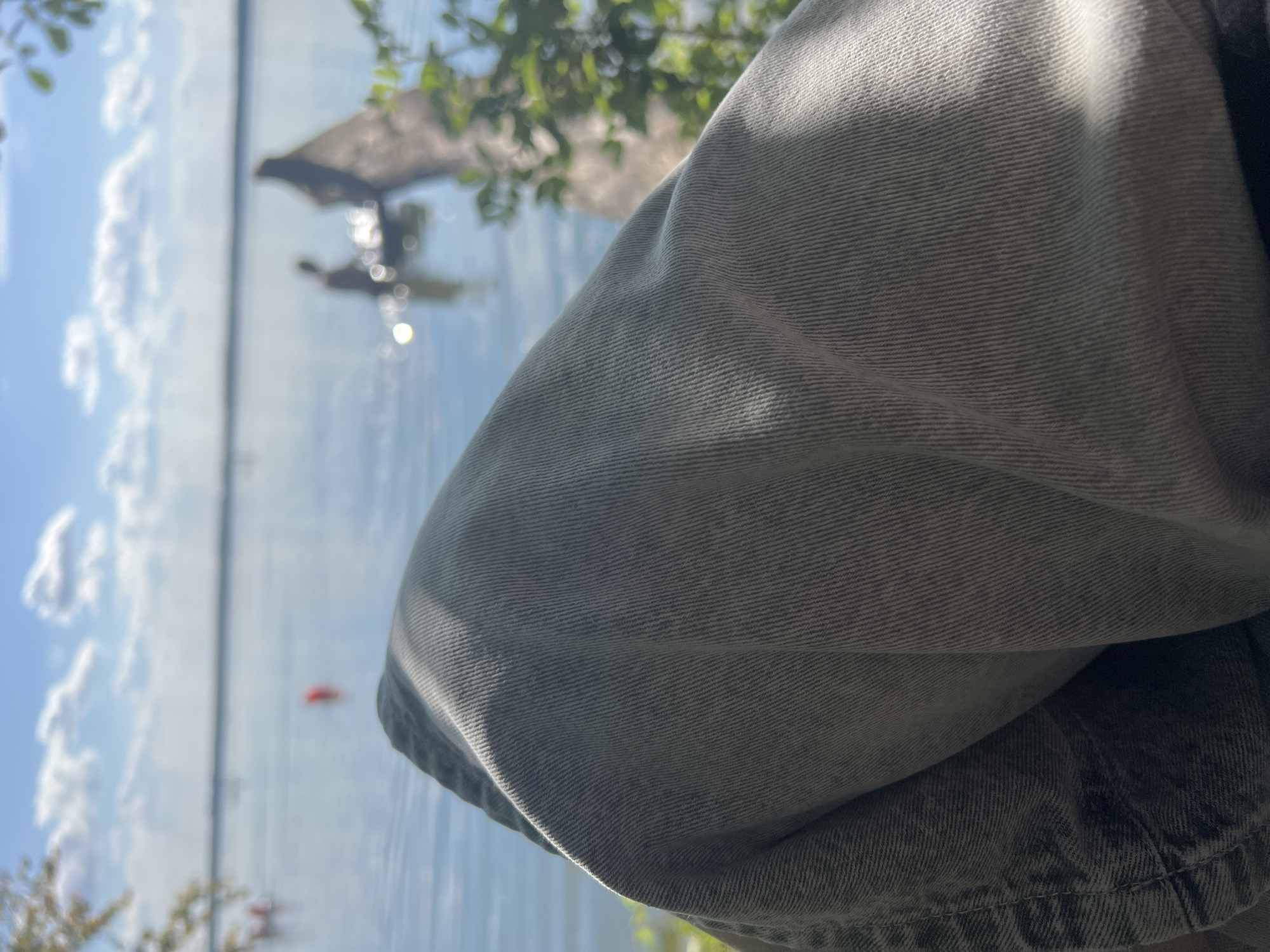 In the city of Romashka, Russia, this image captures a serene lakeside scene. The foreground is dominated by the texture of gray denim pants, suggesting a person is seated and looking out at the water. Beyond the denim, a tranquil lake stretches towards the horizon, reflecting a blue sky dotted with white clouds. A solitary red buoy floats on the water's surface. In the mid-ground, a person is visible in the water, appearing to interact with a fallen tree trunk that juts out from the lake. The scene is illuminated by natural sunlight, casting subtle shadows on the fabric in the foreground. Lush green foliage frames the right side of the image, adding a touch of nature to the composition. The overall atmosphere is one of peaceful recreation by the water.