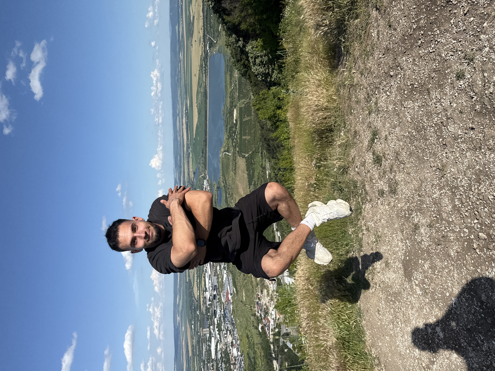 In Shumen, Bulgaria, a man is captured mid-air, appearing to levitate with his legs crossed and arms folded across his chest, against a backdrop of a sprawling city and a calm lake under a bright blue sky dotted with clouds. The man is dressed in a black t-shirt and black shorts, with white sneakers and white socks. He has a short beard and dark hair. The surrounding environment is a grassy, hilly terrain with dry grass in the foreground and lush green vegetation further down the slope. The city of Shumen is visible in the distance, characterized by buildings and infrastructure, and a body of water, likely a lake or reservoir, is prominent in the mid-ground. The overall scene suggests an outdoor recreational setting, possibly a park or a viewpoint overlooking the city and its surroundings.