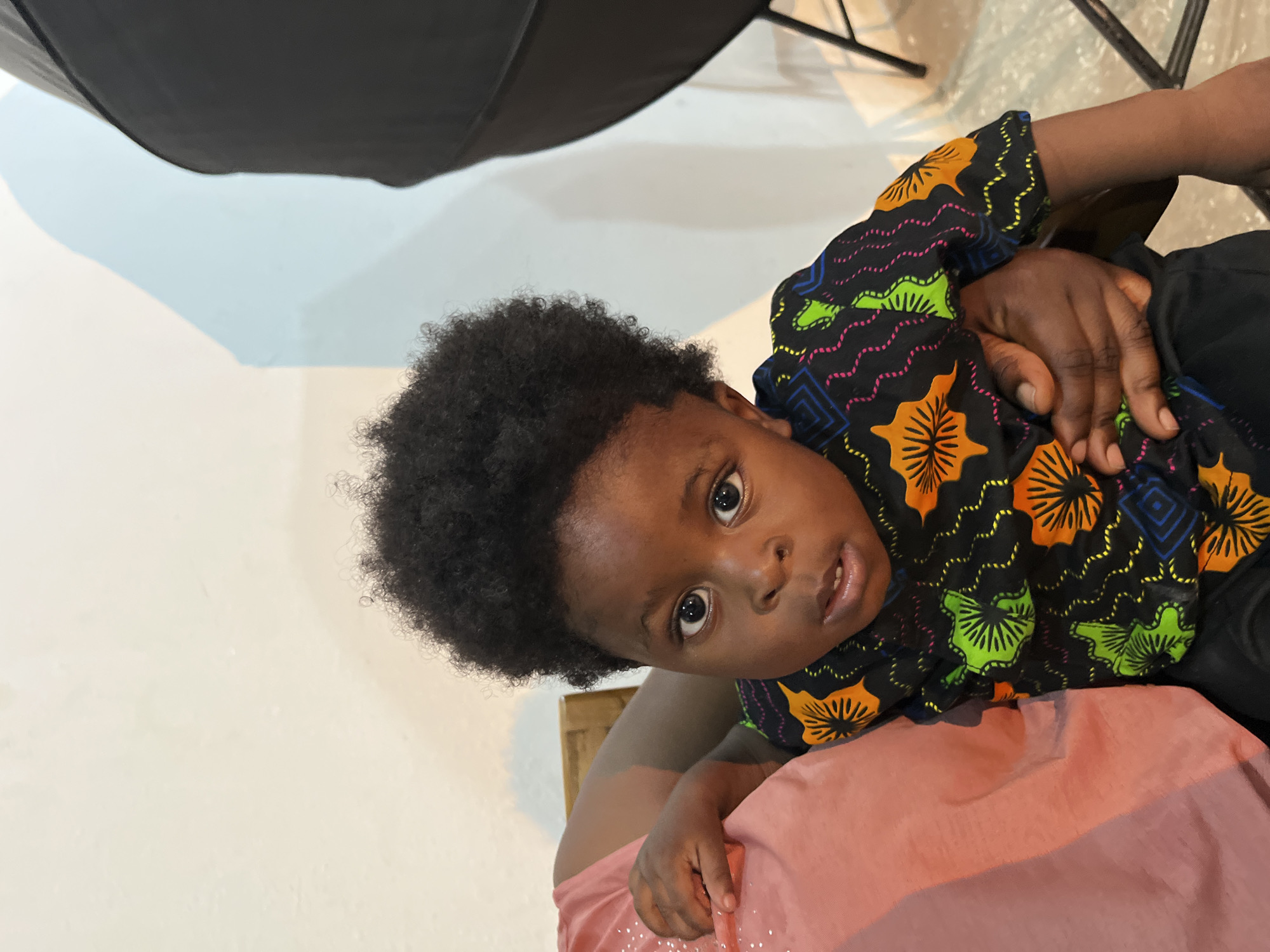 A young Black child with dark, curly hair is being held by an unseen person in a close-up shot. The child is wearing a black t-shirt with a colorful, abstract pattern featuring orange and green circular motifs and wavy lines in pink, yellow, and blue. The child's eyes are open and looking directly at the camera, with a slight pout on their lips. The background is a light-colored wall with a soft blue shadow cast upon it, and a dark, rounded object is visible in the upper right corner, possibly a photographic lighting modifier. The child's left arm is resting on the person holding them, who is wearing a pink garment. The setting appears to be indoors, with the lighting suggesting it could be a home or a studio environment in Gura Top, Nigeria.