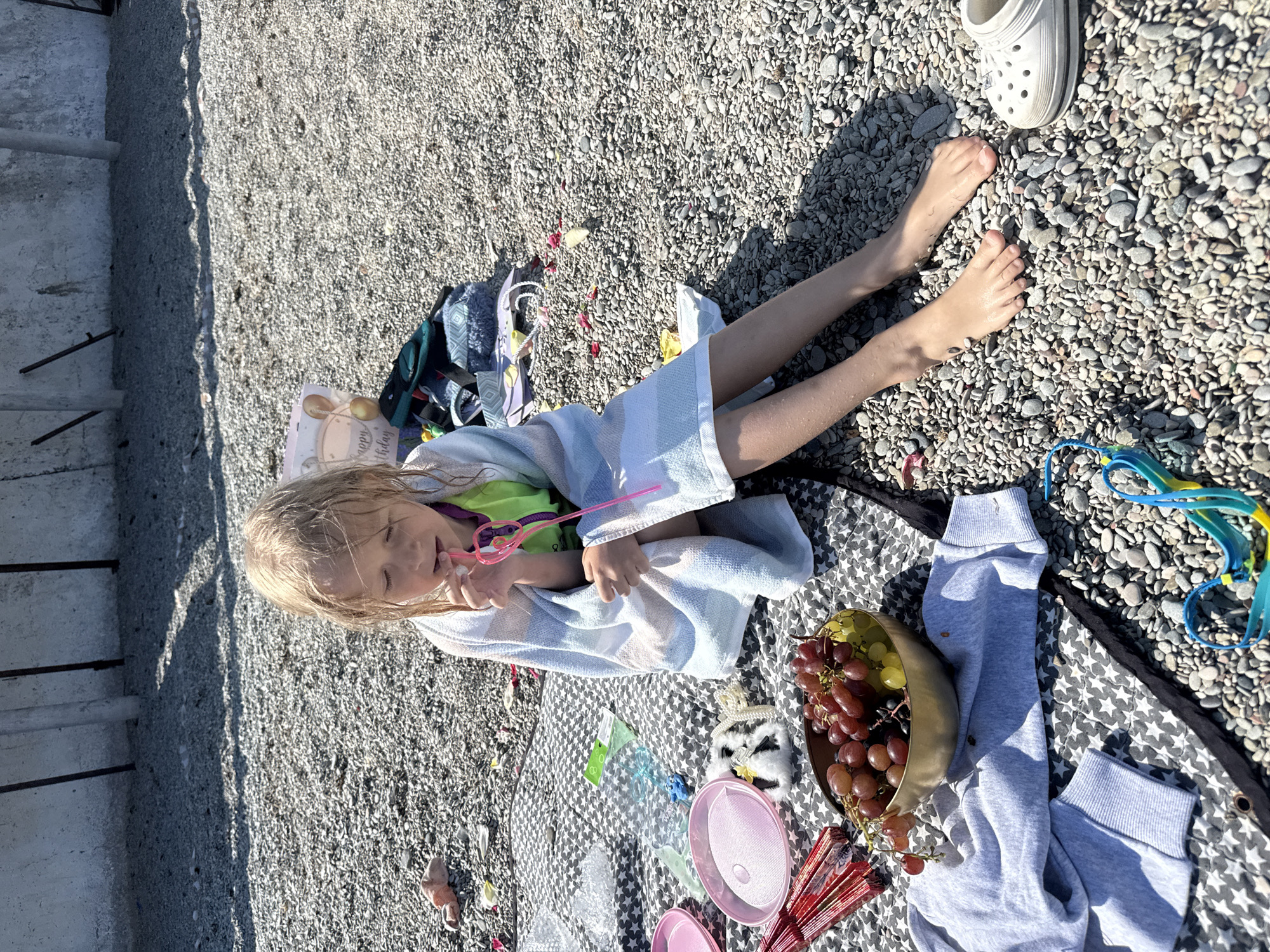 A young child, wrapped in a white towel, sits on a blanket that is spread out on a pebble beach in Unknown, Unknown. The child is fair-haired, with their hair slightly damp, and is holding a pink straw. The blanket is gray and patterned with white stars. Adjacent to the child on the blanket, there's a gold-colored bowl containing grapes, alongside other items like pink toy dishes, a fan, and a stuffed owl. To the right of the child's feet, a white shoe rests on the pebble beach. A pair of blue swimming goggles lies next to the blanket. In the background, a concrete structure is visible to the left of the scene, while the beach itself stretches out towards the top right corner.