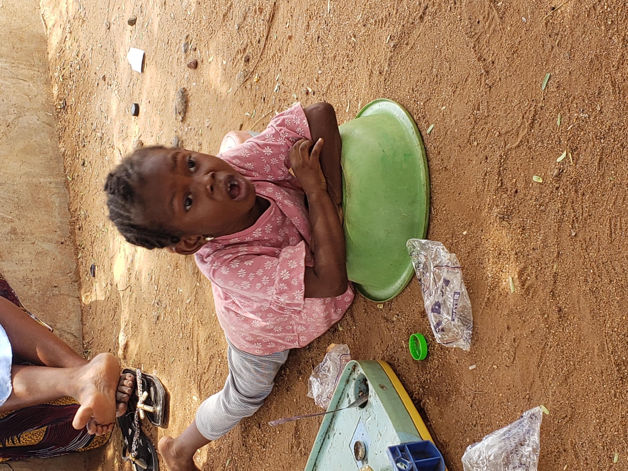 A young child is depicted in an outdoor setting on reddish-brown, dusty ground in Adankolo, Nigeria. The child, likely a girl with dark skin and braided hair, wears a pink short-sleeved top with white floral patterns and light grey striped pants. She is sitting or kneeling, leaning her left arm on a large, bright green plastic basin that is inverted. Her body is turned, and she looks directly towards the viewer with an expressive, open-mouthed gaze, suggesting speech or an immediate reaction.
The ground is dry and littered with small stones, dried leaves, and debris. To the child's lower right, a light blue and yellow plastic toy, possibly a miniature kitchen item or scale, rests on the ground with a small spoon-like utensil on it. A vibrant green plastic bottle cap is also visible nearby, along with two clear plastic sachets. One sachet clearly shows the text "WATER," while another appears to have "RECHARGES" partially visible. In the bottom left corner of the frame, the legs and feet of an adult are visible, wearing black sandals with decorative silver chains, indicating another person is seated close by. The scene is brightly lit by natural light, suggesting it is daytime in a warm environment.