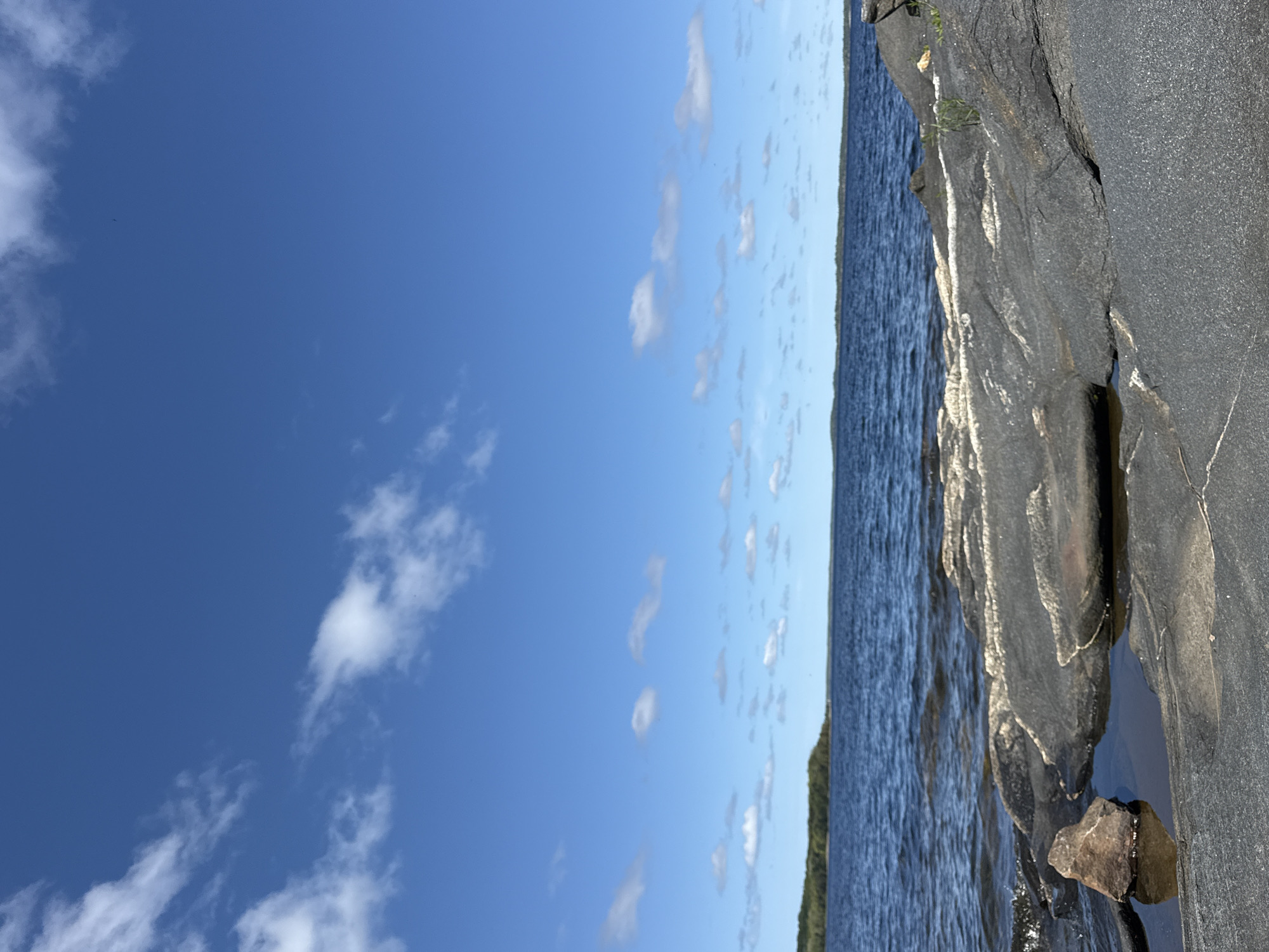 The image presents a view of a body of water meeting a rocky shoreline under a blue sky. The sky, occupying the left portion of the frame, is clear with scattered white clouds. The water, with a textured surface indicating possible ripples or waves, extends towards the horizon where it meets a strip of greenery. The right side of the image is dominated by large, dark rocks with visible layers and crevices, suggesting natural formations. The location appears to be a natural environment, possibly a lake or coastal area, but lacks specific identifiable landmarks to determine the exact location.