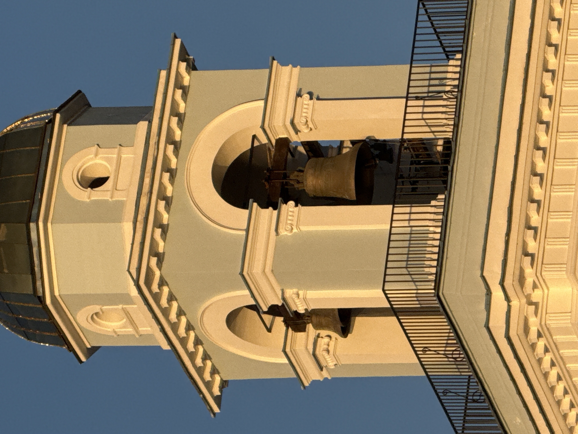The image shows a close-up view of a bell tower on a clear, blue-sky day, likely in Votkinsk, Russia. The tower is painted a pale blue with white architectural details. Two large, bronze bells hang within arched openings in the tower. A dark metal railing is visible on a balcony level of the tower. The lighting suggests it is either early morning or late afternoon, casting warm, golden light on the building. No people are visible in the image.