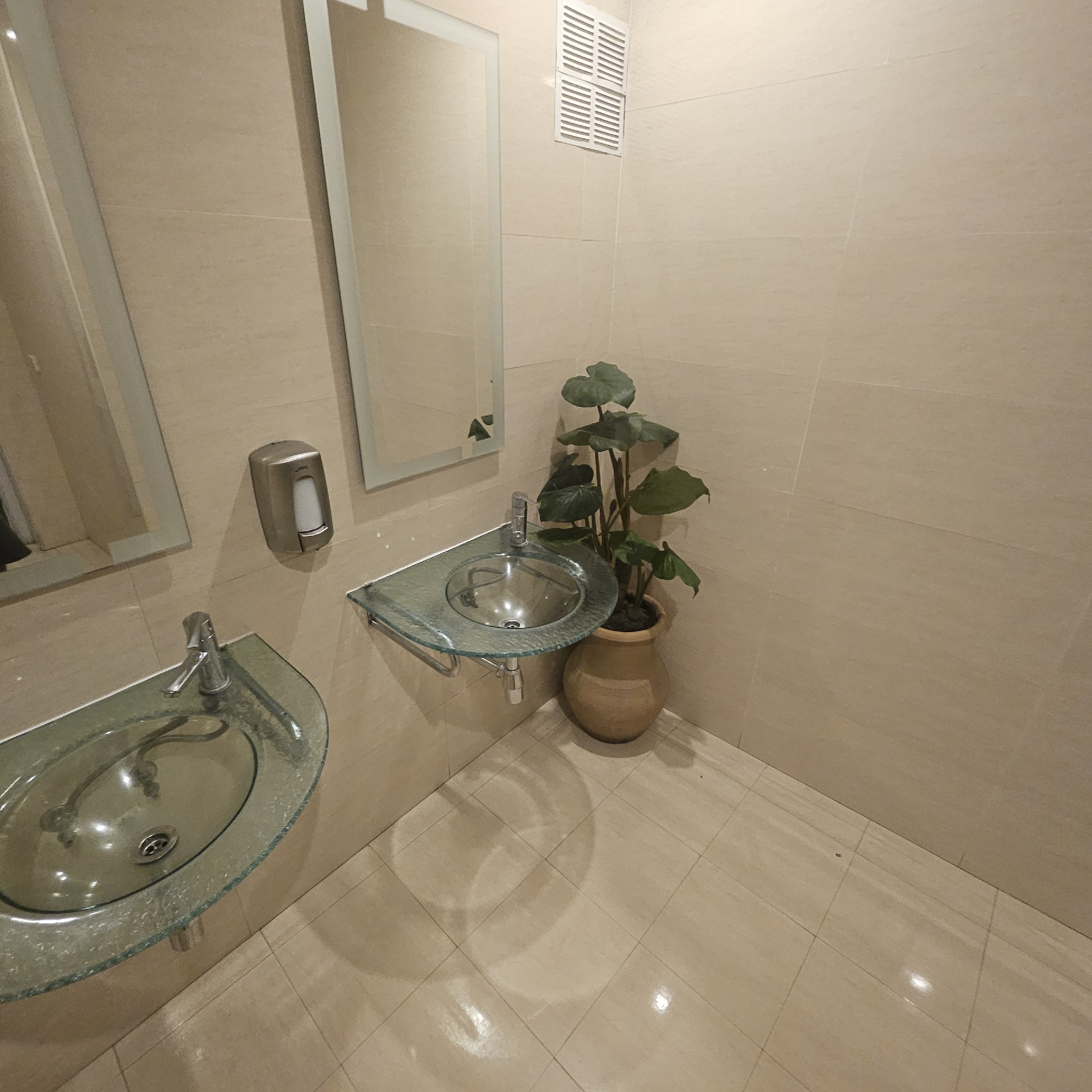 The image depicts a clean, modern public restroom in Almadrones, Spain. The walls and floor are tiled with large, light-colored square tiles, creating a bright and hygienic atmosphere. There are two sinks visible. The sink on the left is a semi-circular glass basin with a chrome faucet, mounted on the wall with visible plumbing underneath. Above this sink, a chrome soap dispenser is mounted on the wall. To the right, another, smaller, semi-circular glass sink with a chrome faucet is also wall-mounted. Adjacent to this sink, a large potted plant with broad green leaves adds a touch of nature to the space. In the upper right portion of the image, a white air vent is installed in the tiled wall. A large rectangular mirror hangs above the right sink, reflecting a portion of the room. The lighting appears to be artificial, casting a warm glow on the surfaces. No individuals are present in the image.