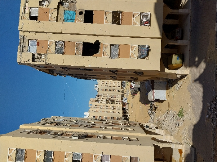 The image depicts a devastated urban landscape in Khan Yunis, Palestinian Territory, characterized by heavily damaged multi-story residential buildings under a clear blue sky. Two prominent, similarly constructed light beige buildings with brown decorative window surrounds frame the foreground, exhibiting extensive structural damage. This damage includes numerous large holes in exterior walls, missing windows, shattered glass, and exposed interiors across multiple floors. Some window openings are covered with various fabrics, including a blue tarp on the right building and white or dark cloths elsewhere.
Between these foreground structures lies a wide, unpaved dirt and rubble-strewn area. Piles of concrete debris and scattered construction materials are visible on the ground. Several makeshift structures, including a white tent-like shelter with a brown roof, are erected in the open space. Further in the background, more residential buildings, also appearing damaged, line the horizon. Laundry hangs on clotheslines near other informal structures in the middle ground.
The ground floor of the right foreground building features an open arcade supported by pillars, under which a yellow, tarp-covered object (possibly a vehicle) and a silver sedan are parked. The ground beneath the left building's arcade also shows debris. Overhead, a thin wire or cable stretches horizontally between the upper floors of the two foreground buildings. Shadows from unseen structures in the immediate foreground and the buildings themselves are cast across the dusty ground. No people are explicitly visible in the scene.