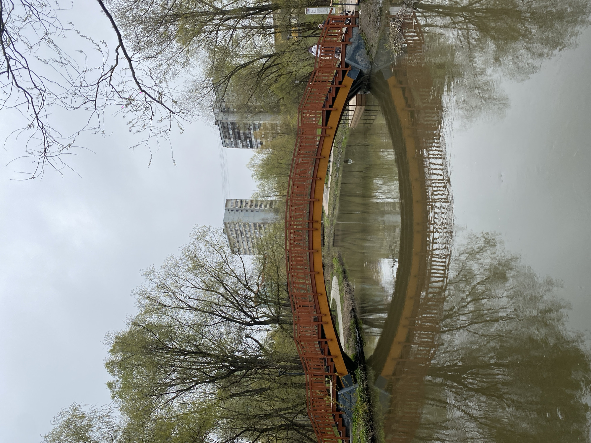 The image depicts a curved pedestrian bridge spanning a body of water, likely a pond or small lake, in a park-like setting. The bridge has a brown railing and a yellow structure. The water reflects the bridge, trees, and nearby buildings, creating a symmetrical effect. Bare tree branches and trees with budding leaves are visible along the banks of the water. In the background, there are apartment buildings, suggesting this location is in a city. The sky is overcast, indicating it is likely daytime but under cloudy conditions. Based on the appearance of the buildings and vegetation, the location is likely the Serebryany Bor in Moscow, Russia. There is one person on the bridge.