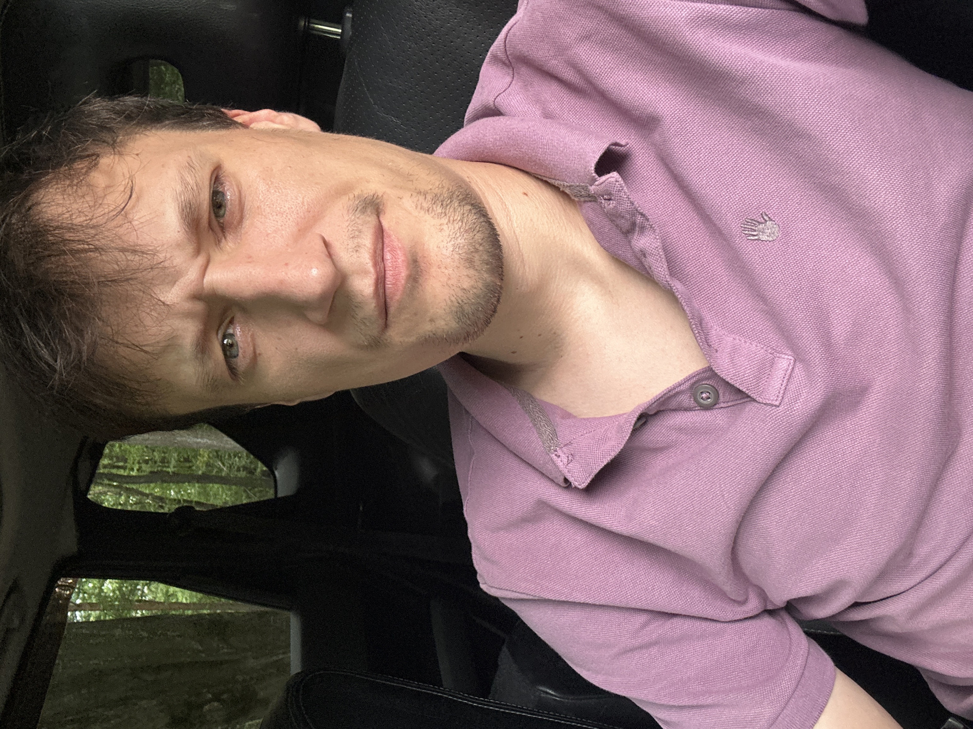 A man is captured in a close-up, selfie-style portrait inside a vehicle. He is seated in the driver's seat, with the car's interior visible behind him, including the dark seats and the windows showing a blurred, green forest environment. The man has short, dark hair and is wearing a light purple polo shirt with a small embroidered hand logo on the left side of the chest. His facial expression is one of mild concern or skepticism, with furrowed brows and slightly narrowed eyes. He has light stubble on his chin and upper lip. The lighting suggests it is daytime, possibly overcast, given the soft illumination. The image was taken in Shatava, Ukraine.
