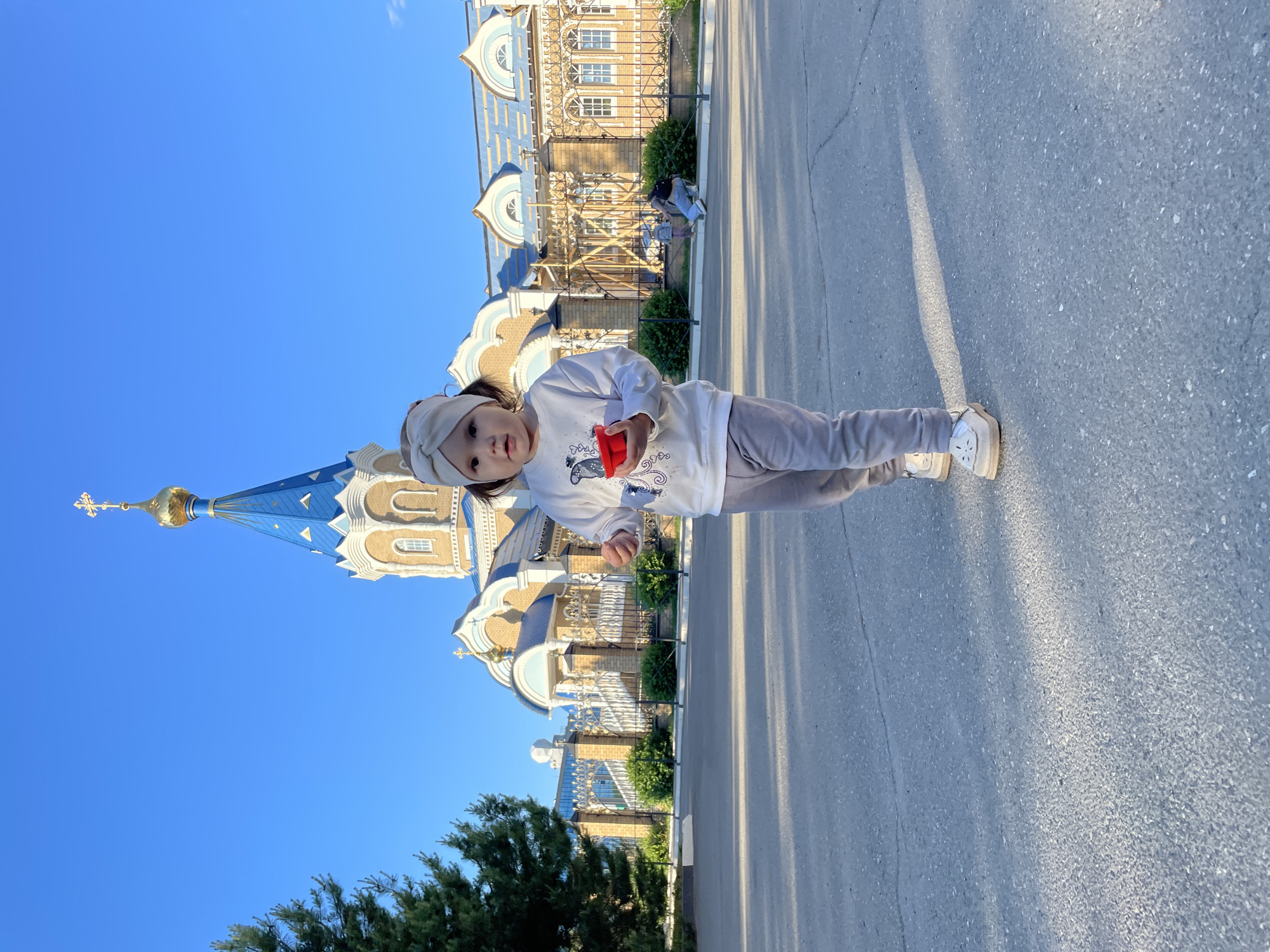 A young child, wearing a white sweatshirt with a graphic print, gray pants, and white shoes, walks towards the camera on an asphalt path. The child holds a small red object in their right hand. In the background, a church with a blue roof and golden dome stands under a clear blue sky. A fence surrounds the church, and some trees are visible to the left. The church is located in Izhevsk, Russia. The lighting suggests it is daytime, with shadows cast on the path.