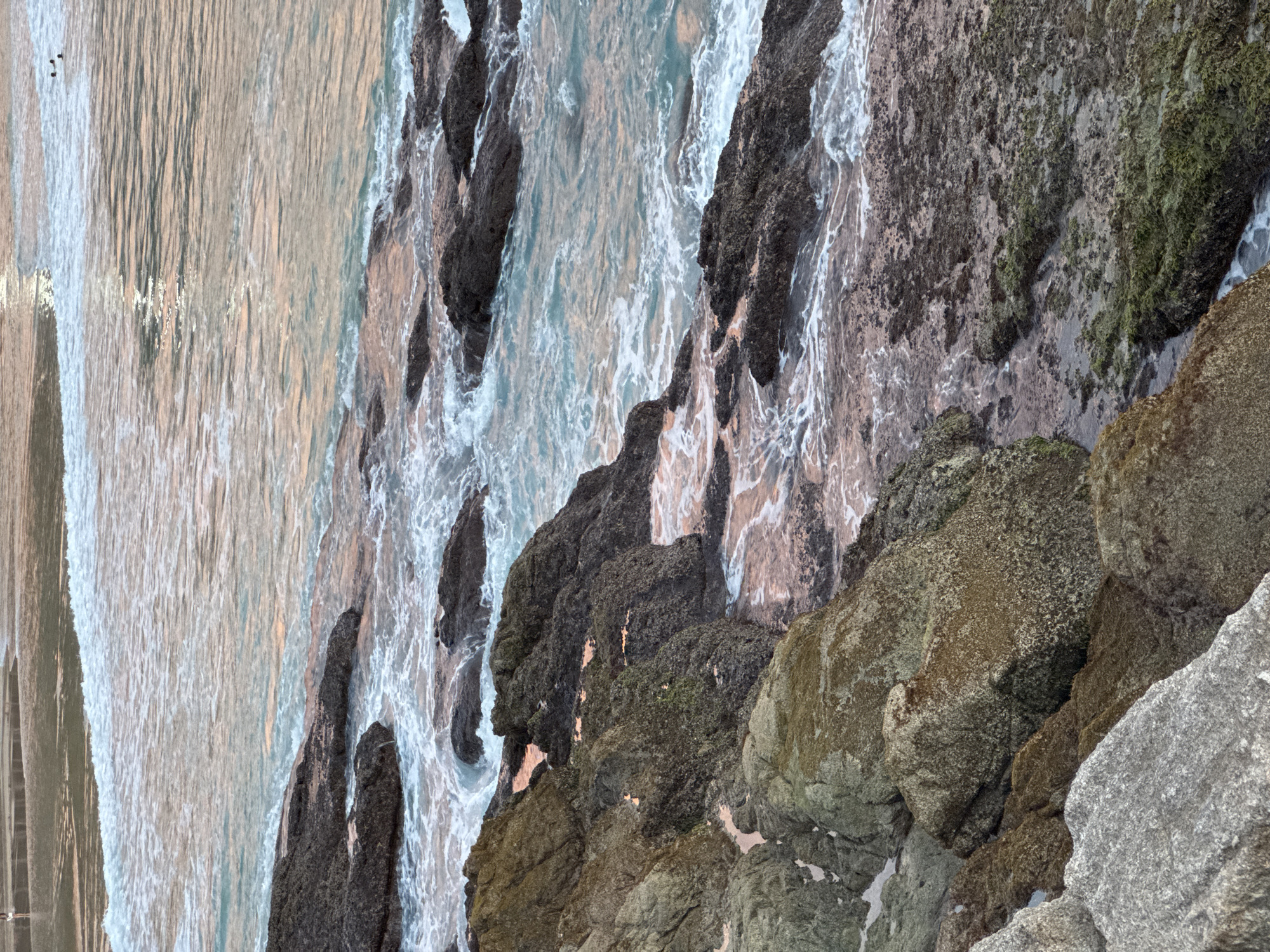 An eye-level, high-angle medium shot captures the rocky coastline of Choeng Thale, Thailand, at what appears to be dusk or dawn. Dark, jagged rocks, some covered in moss and lichen, jut out from the water, creating a textured foreground and midground. The sea, a blend of turquoise and pastel pink hues, gently laps against the rocks, forming white frothy waves. In the background, a shallow stretch of water reflects the soft light, and two indistinct figures can be seen wading or swimming in the distance. The overall scene conveys a sense of natural beauty and tranquility, characteristic of a tropical beach setting.