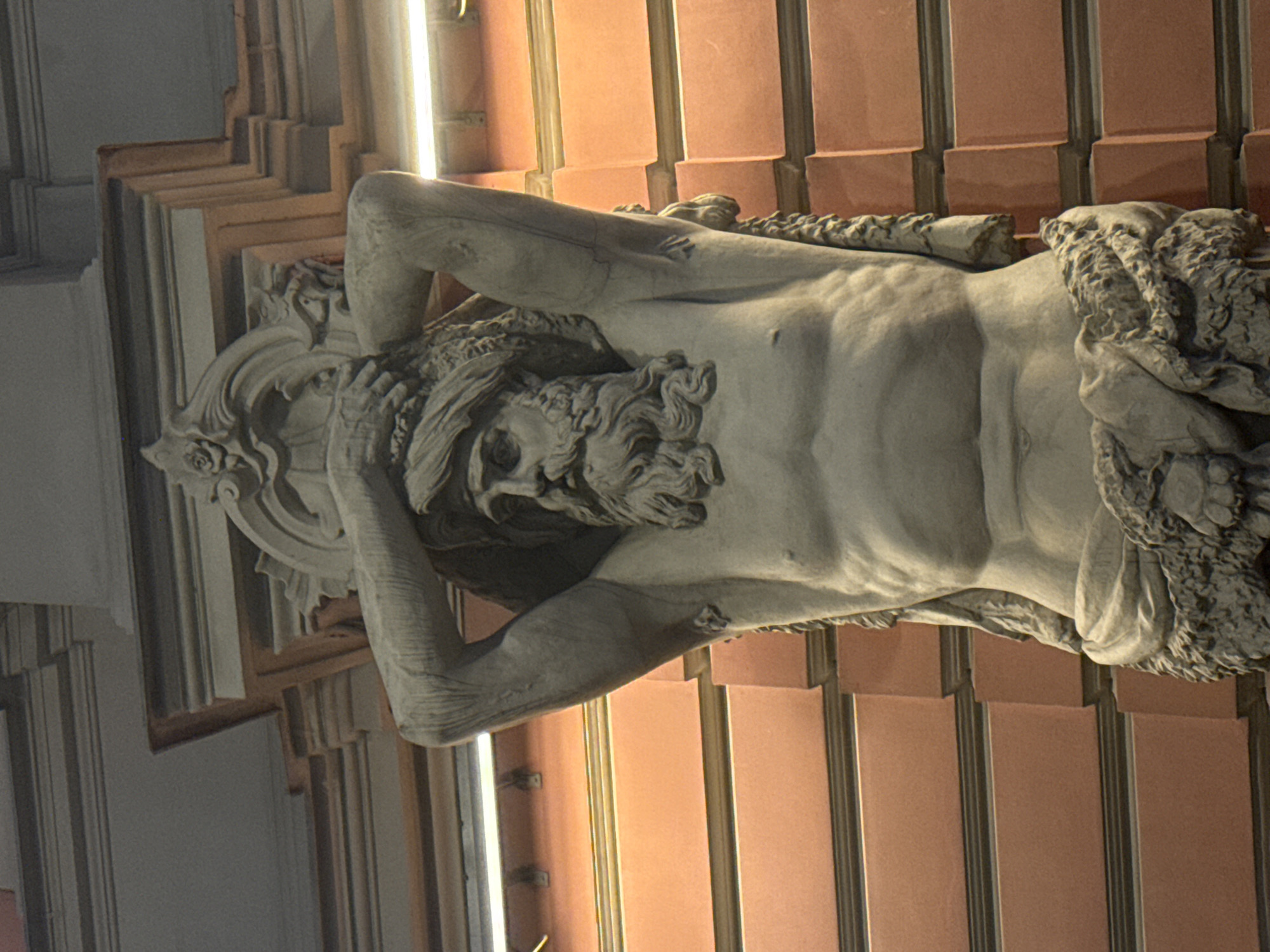 This is a close-up, eye-level shot of a stone sculpture of a muscular, bearded male figure carrying an ornate architectural element on his shoulders. The sculpture is a caryatid, a sculpted female figure used as an architectural support, but in this case, it is a male figure, also known as an Atlas or telamon. The figure is depicted from the torso up, with his arms raised to support the weight above his head. He has a stern expression on his face, with his eyes open and his mouth closed. He is wearing a head covering and a draped garment around his waist. The background consists of a salmon-colored brick wall with horizontal banding, and what appear to be fluorescent lights running horizontally above the sculpture. The sculpture is likely located on the exterior of a building in Saint Petersburg, Russia.