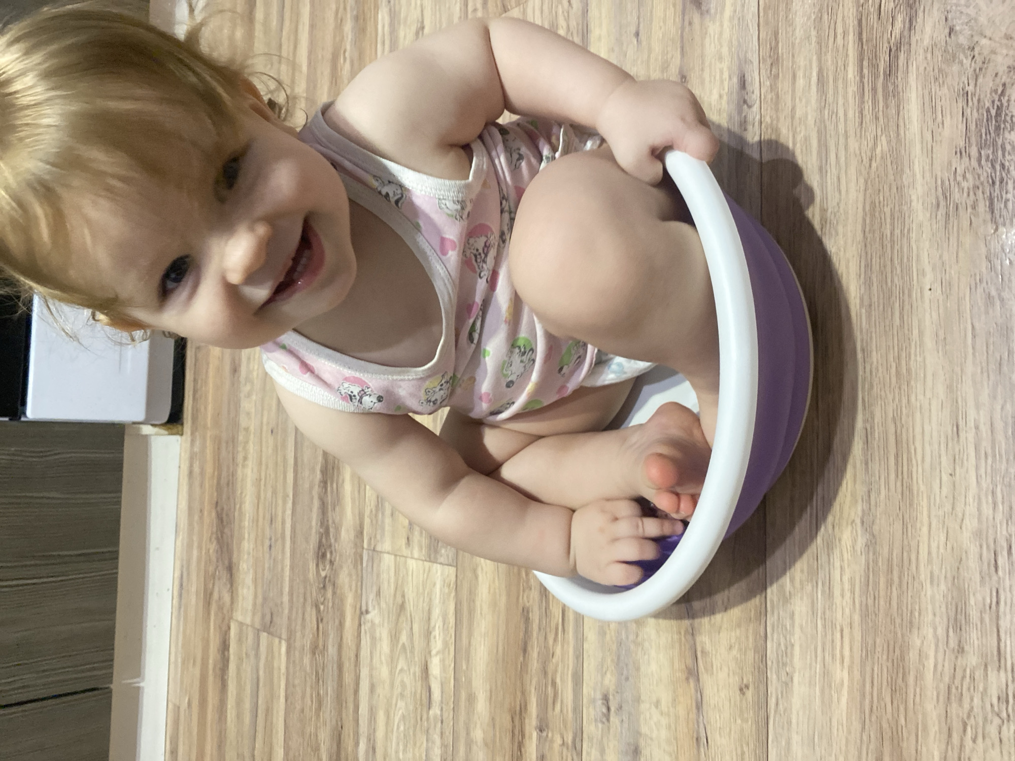 A baby girl is sitting in a purple bowl on a wooden floor indoors. She is wearing a pink and white printed tank top. She is smiling and looking up. A white cabinet is visible in the background to the left.
