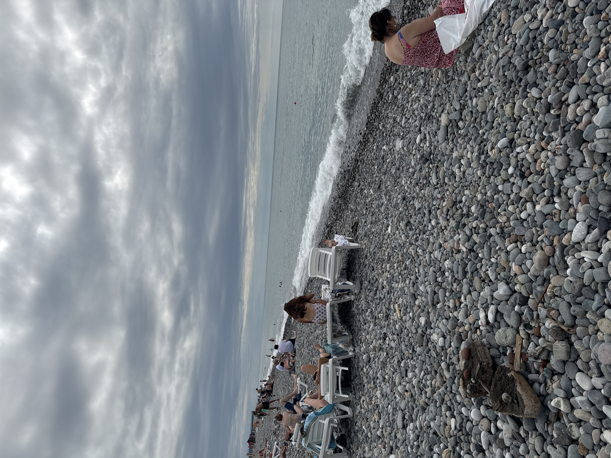 The image depicts a crowded pebble beach in Batumi, Georgia, under an overcast sky. The sea, with moderate waves, stretches to the horizon. Several people are visible on the beach, some reclining on lounge chairs, others sitting or walking. On the right side of the frame, a woman in a red floral dress is seated on the pebbles, a white bag beside her. In the foreground, numerous grey and dark pebbles cover the beach. Further down the shore, a line of lounge chairs and people are situated closer to the water. The overall scene suggests a typical summer day at a popular beach destination.