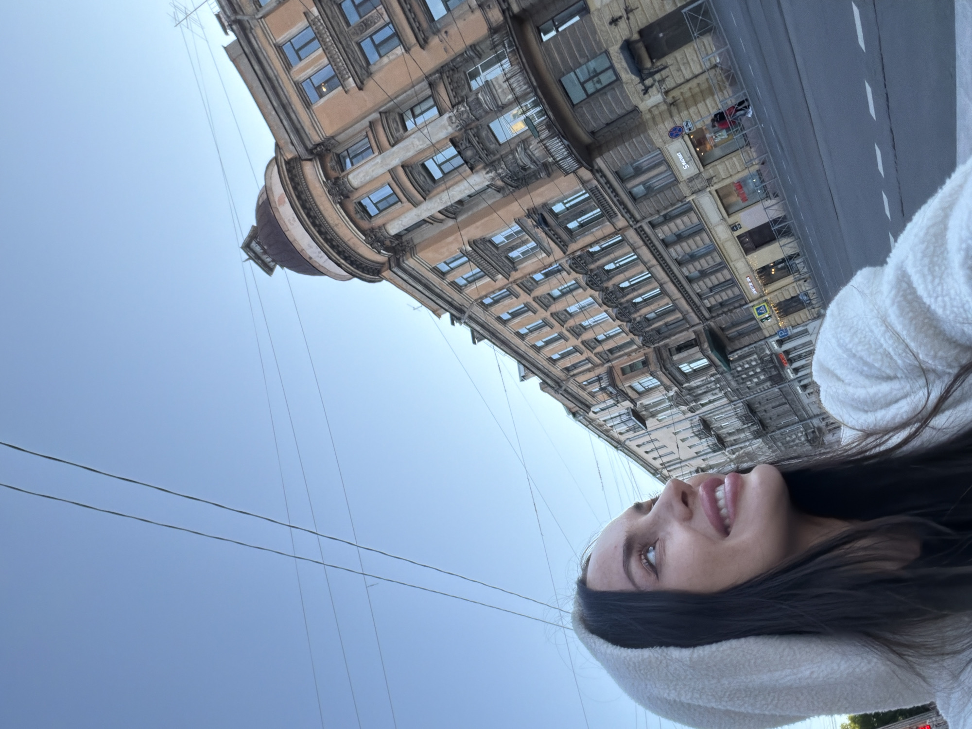 The image captures a low-angle selfie of a young woman with dark hair, wearing a white hooded sweatshirt. She is looking upwards and to her right, with a smile. Behind her, a street scene unfolds with an ornate, multi-story building characteristic of Saint Petersburg, Russia. The building features a prominent dome and detailed architectural elements, with several windows and balconies visible. The street below shows a road with lane markings and a sidewalk lined with shops. Overhead, power lines crisscross the sky. The overall impression is an urban setting in Saint Petersburg, Russia, during daylight, with a focus on the woman and the architectural backdrop.